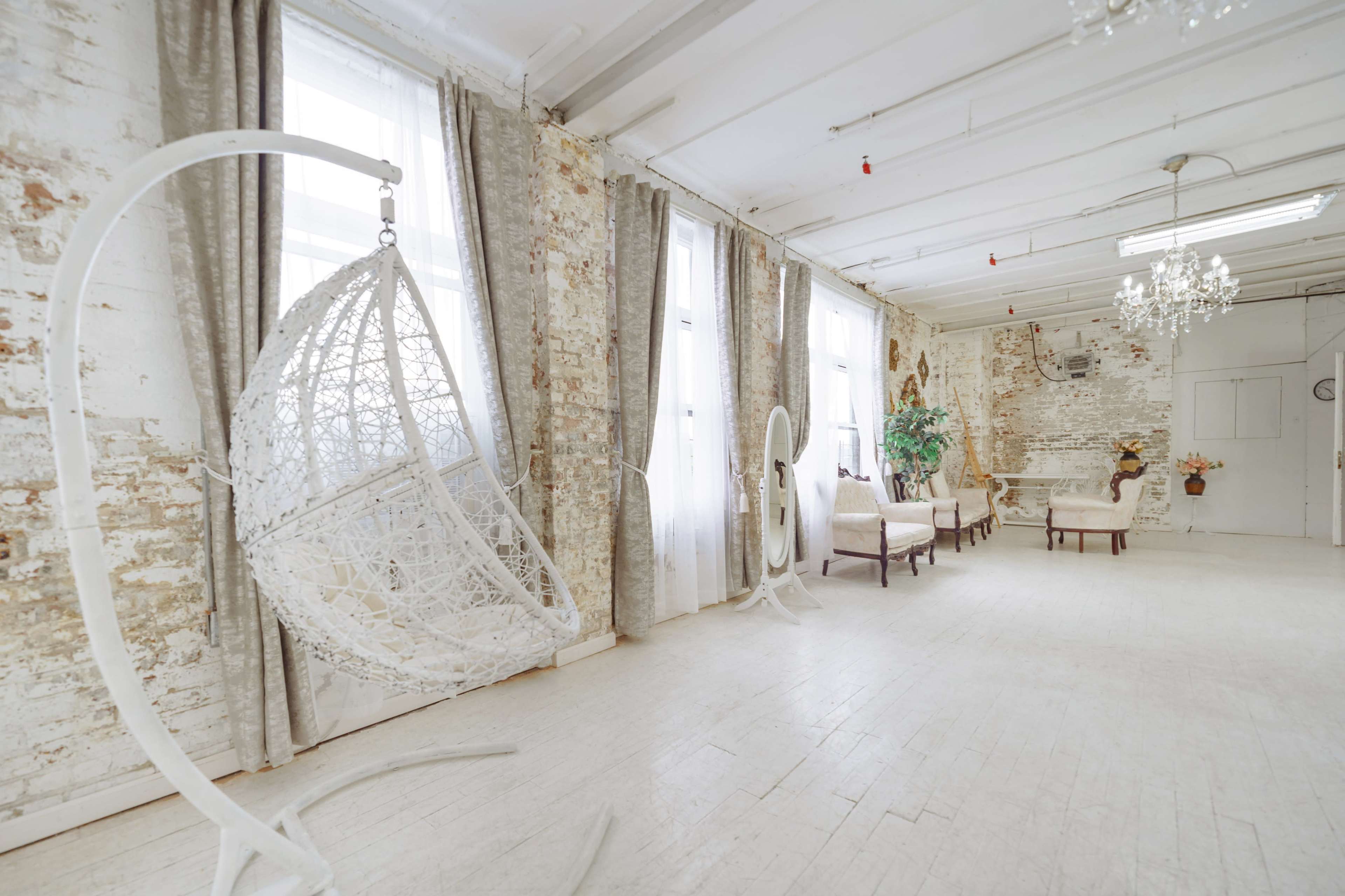 A spacious, lightly decorated room with white walls, large windows, and a hanging basket chair, along with vintage-style furniture arranged along one side.