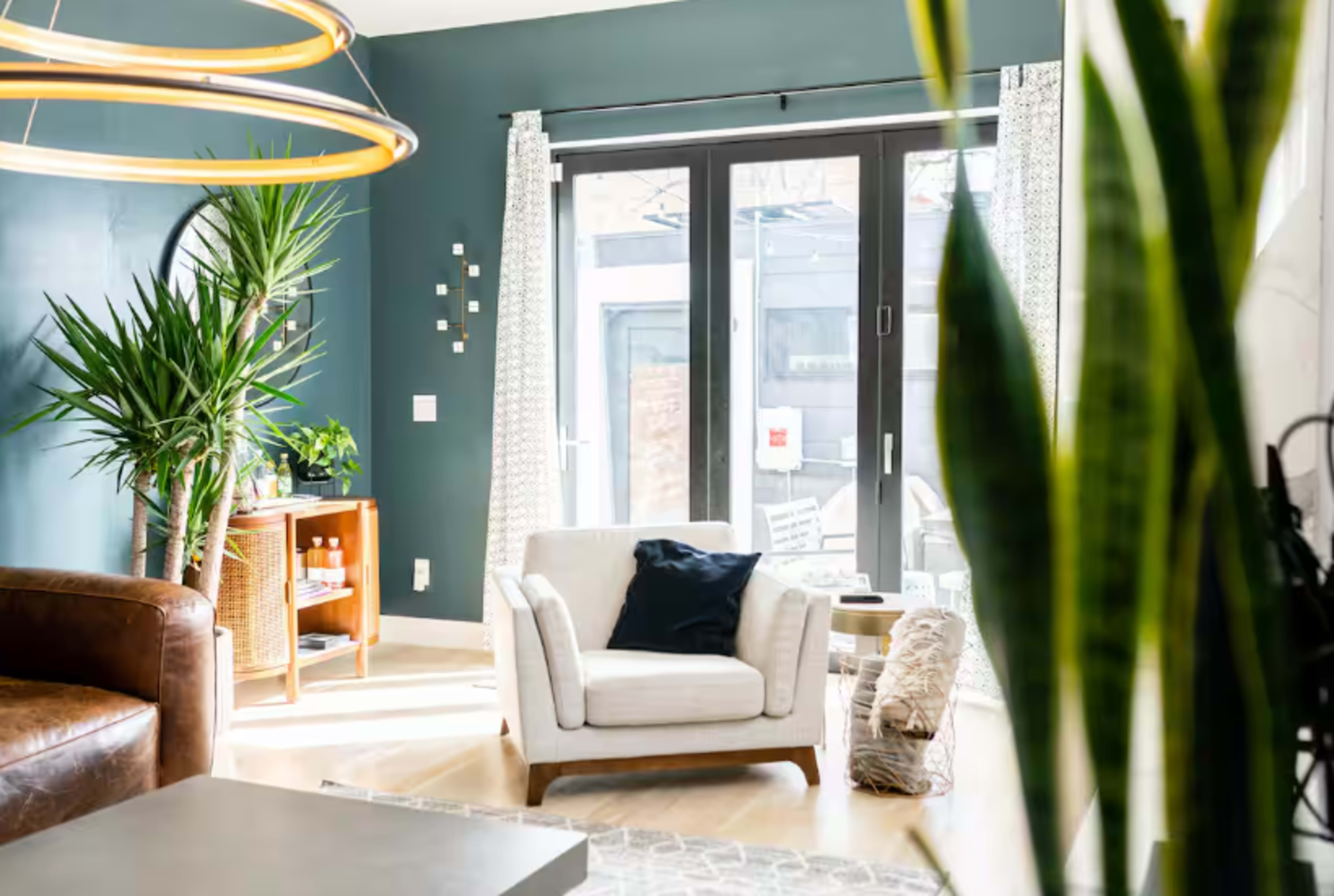 The image shows a well-lit living room featuring a white armchair, a wooden cabinet, and large windows with outdoor views, all set against a teal wall.