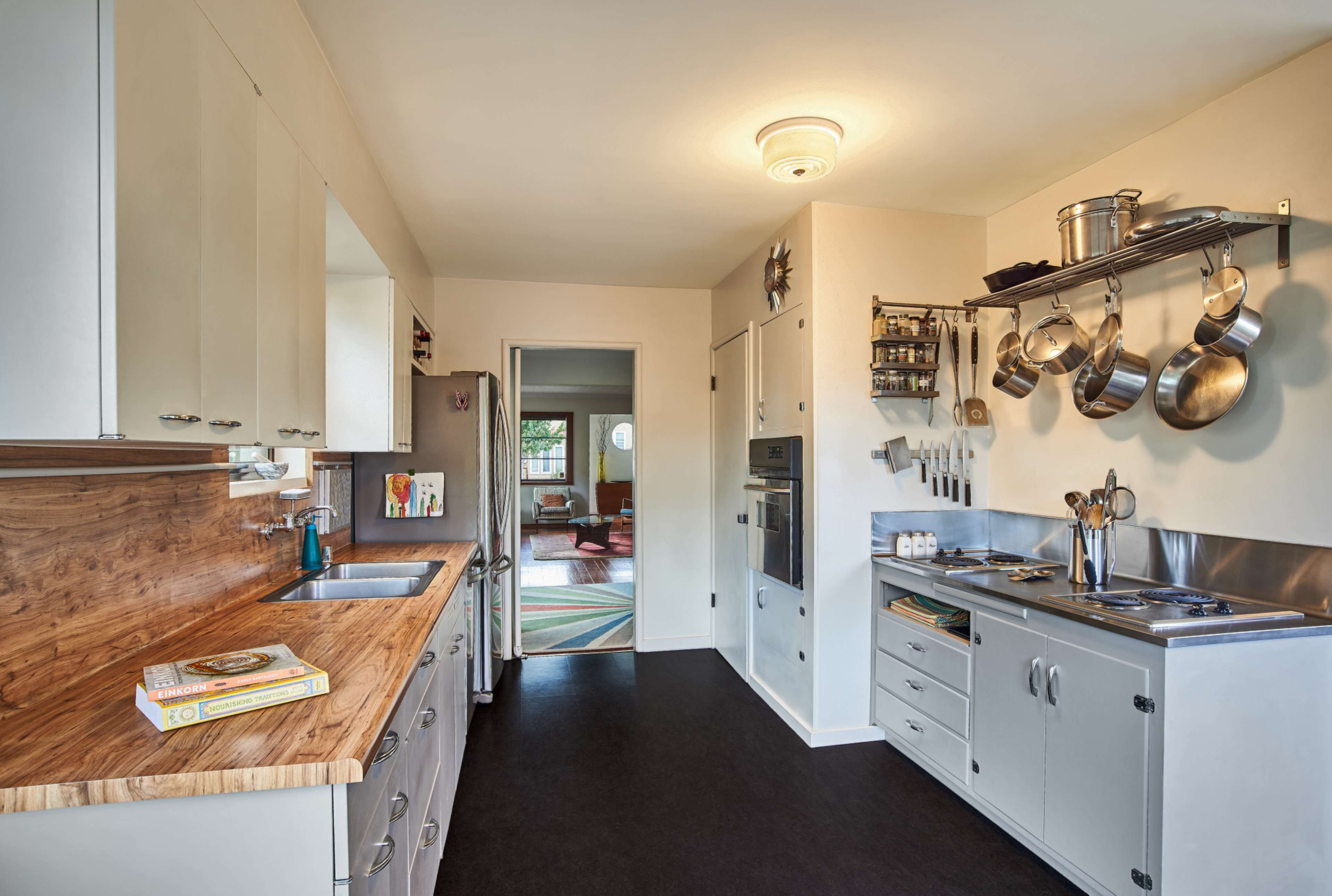 A modern kitchen features stainless steel appliances, a wooden countertop, and hanging pots on a wall rack, with an adjacent living area visible through an open doorway.