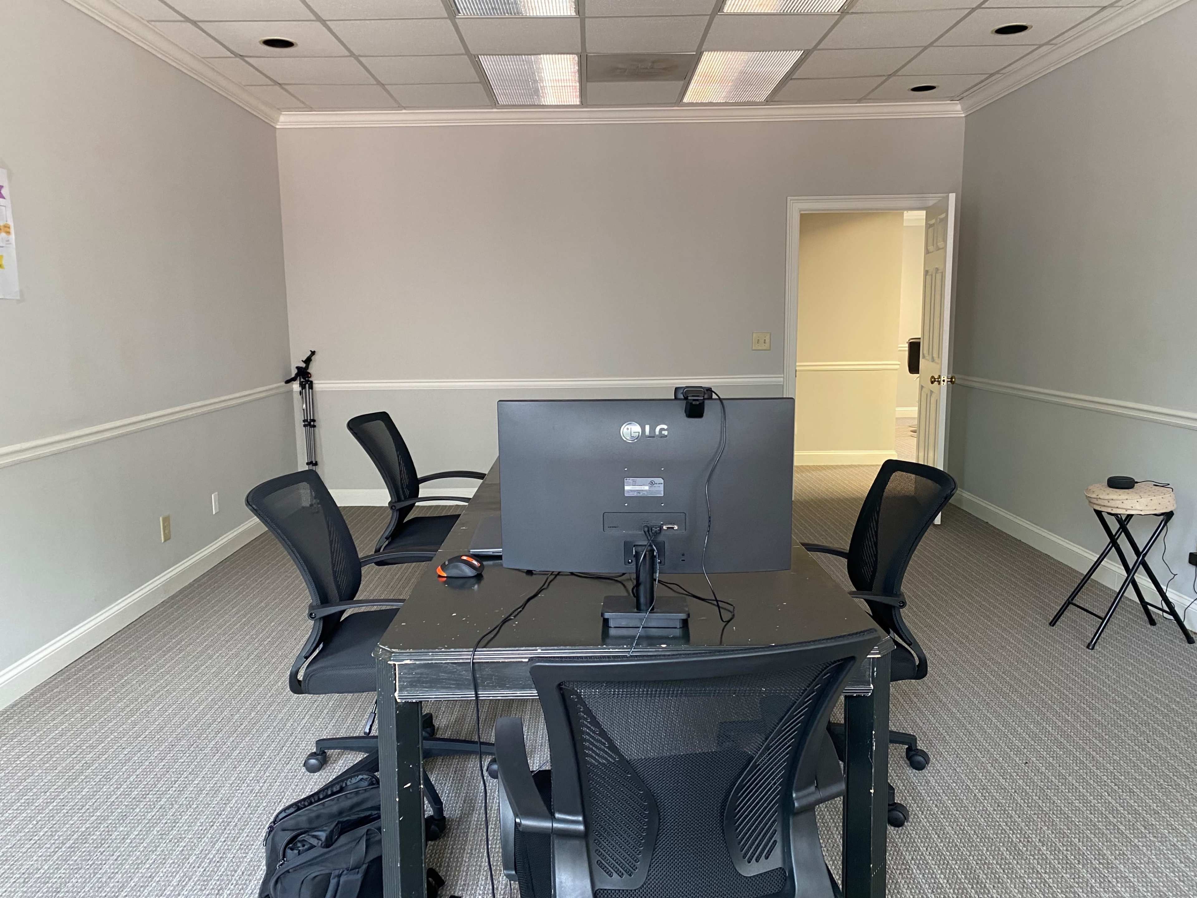 An empty office space features a table with a computer monitor and four black chairs, all set against a neutral-colored wall.