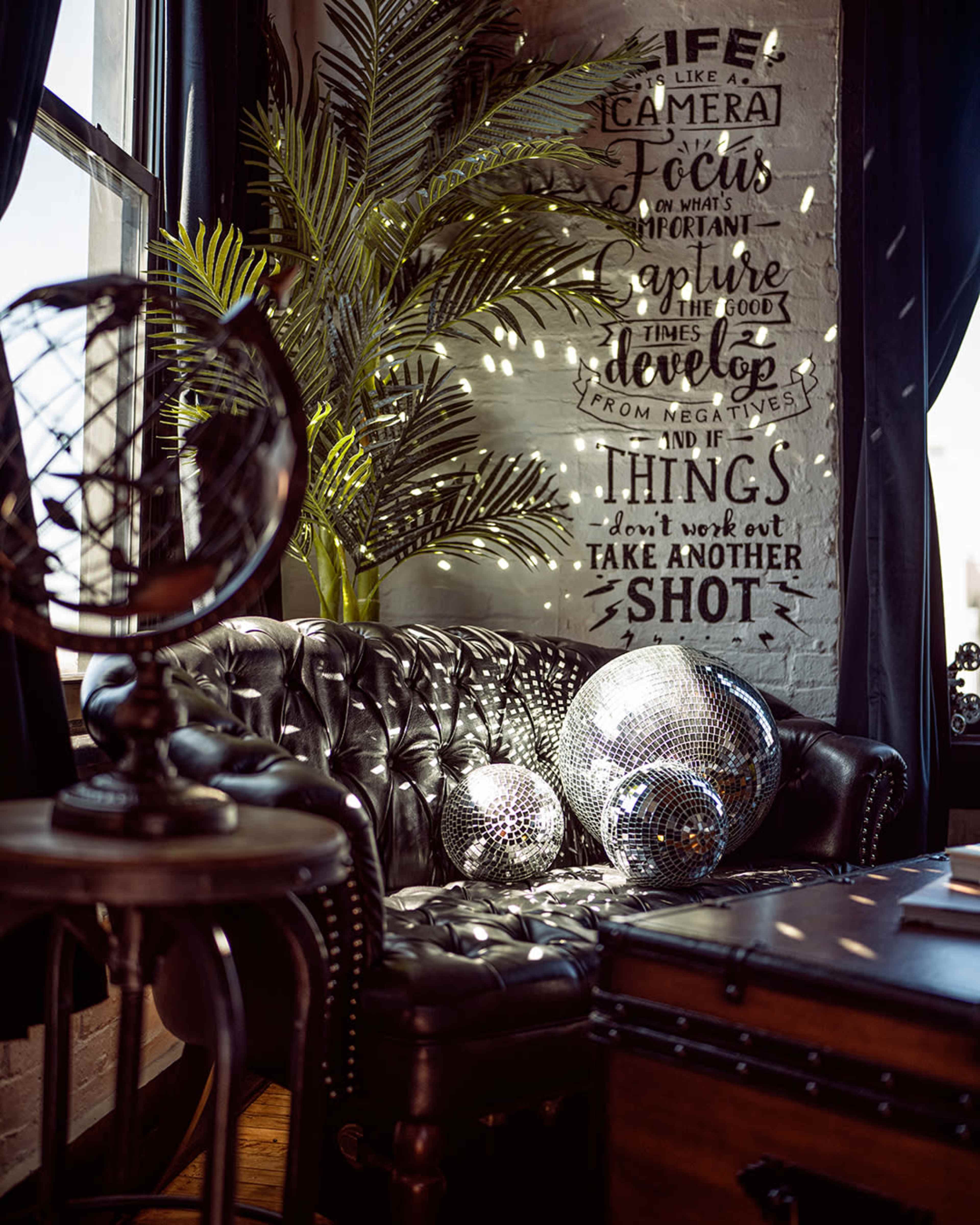 The image features a cozy corner with a black leather sofa, a globe, and reflective disco balls, set against a wall with inspirational text and green foliage.