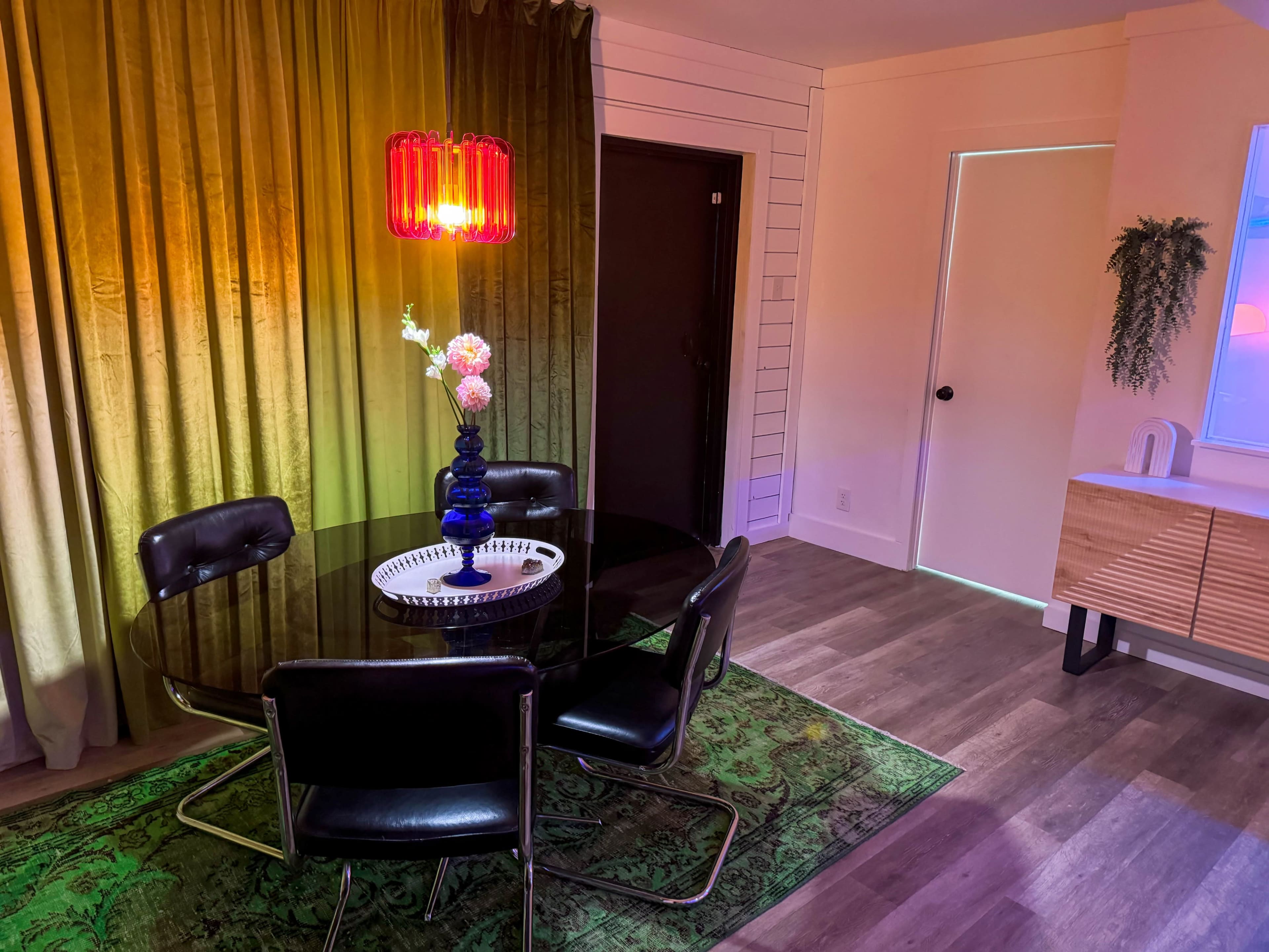 A round black dining table with four chairs is set in a room featuring a green rug, colorful lighting, and a large curtain backdrop.