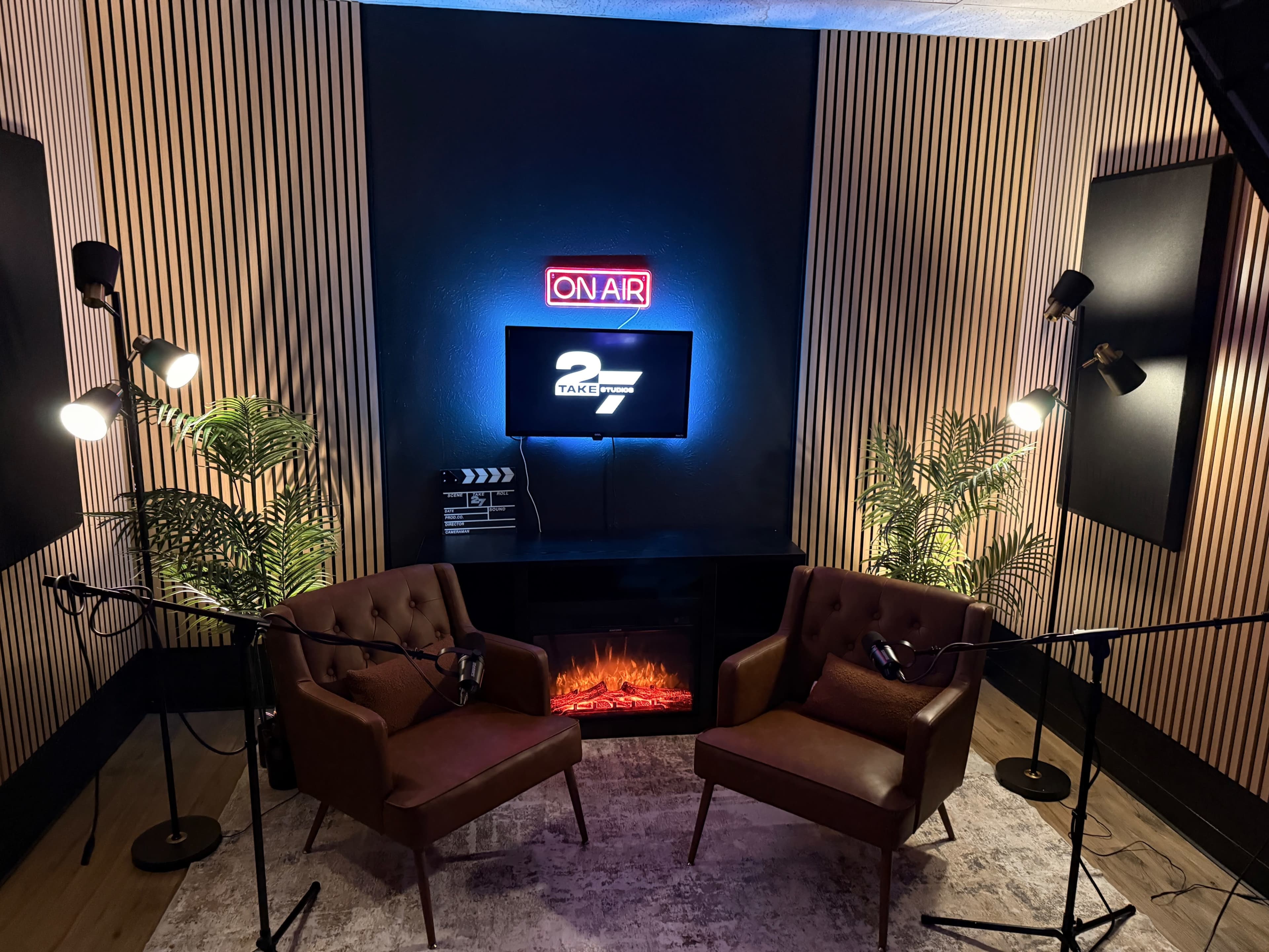 The image shows a cozy recording studio with two brown chairs facing a neon "ON AIR" sign, along with floor lamps, a television screen, and potted plants.