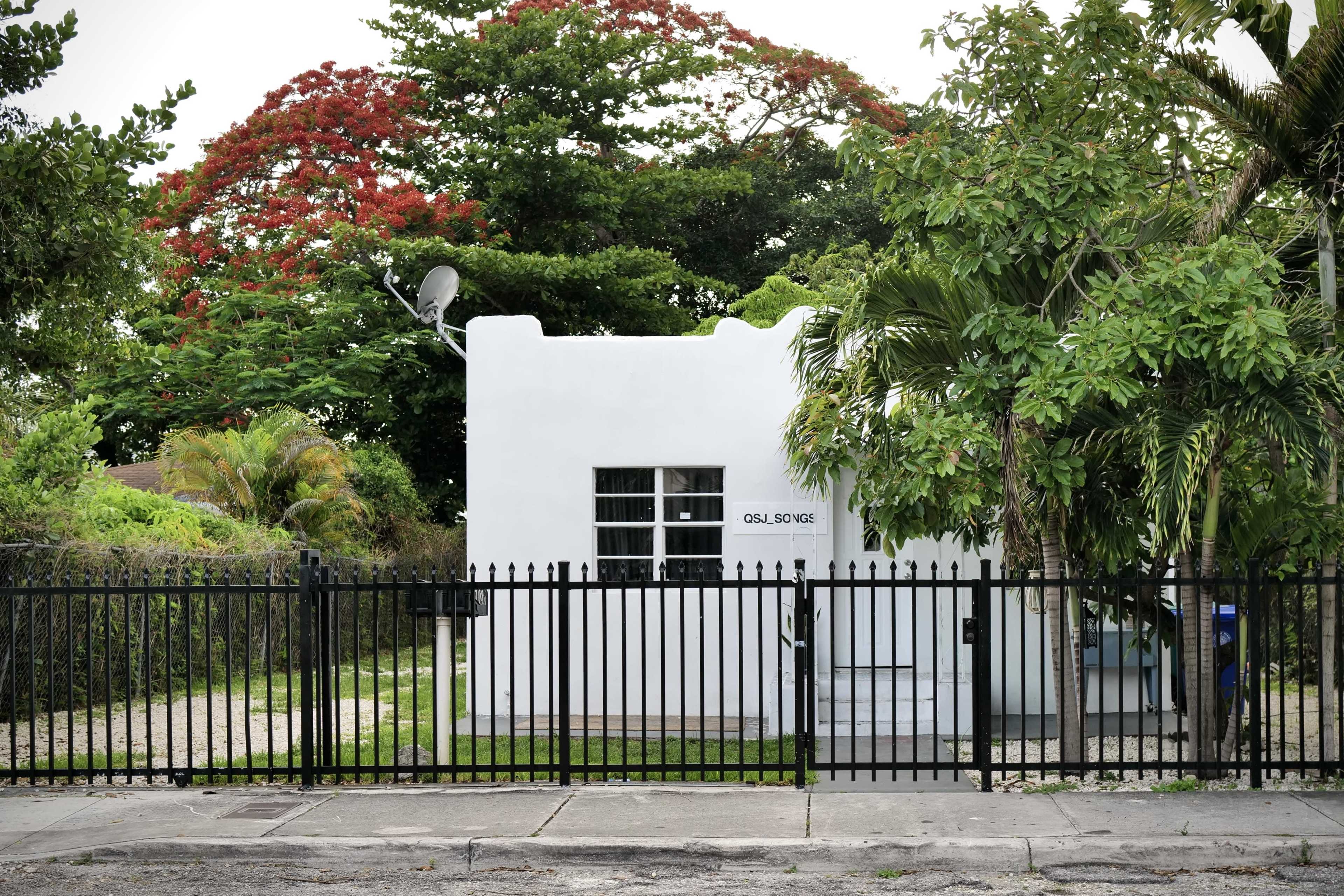 A small white building with a gated fence is surrounded by lush greenery and blooming trees.