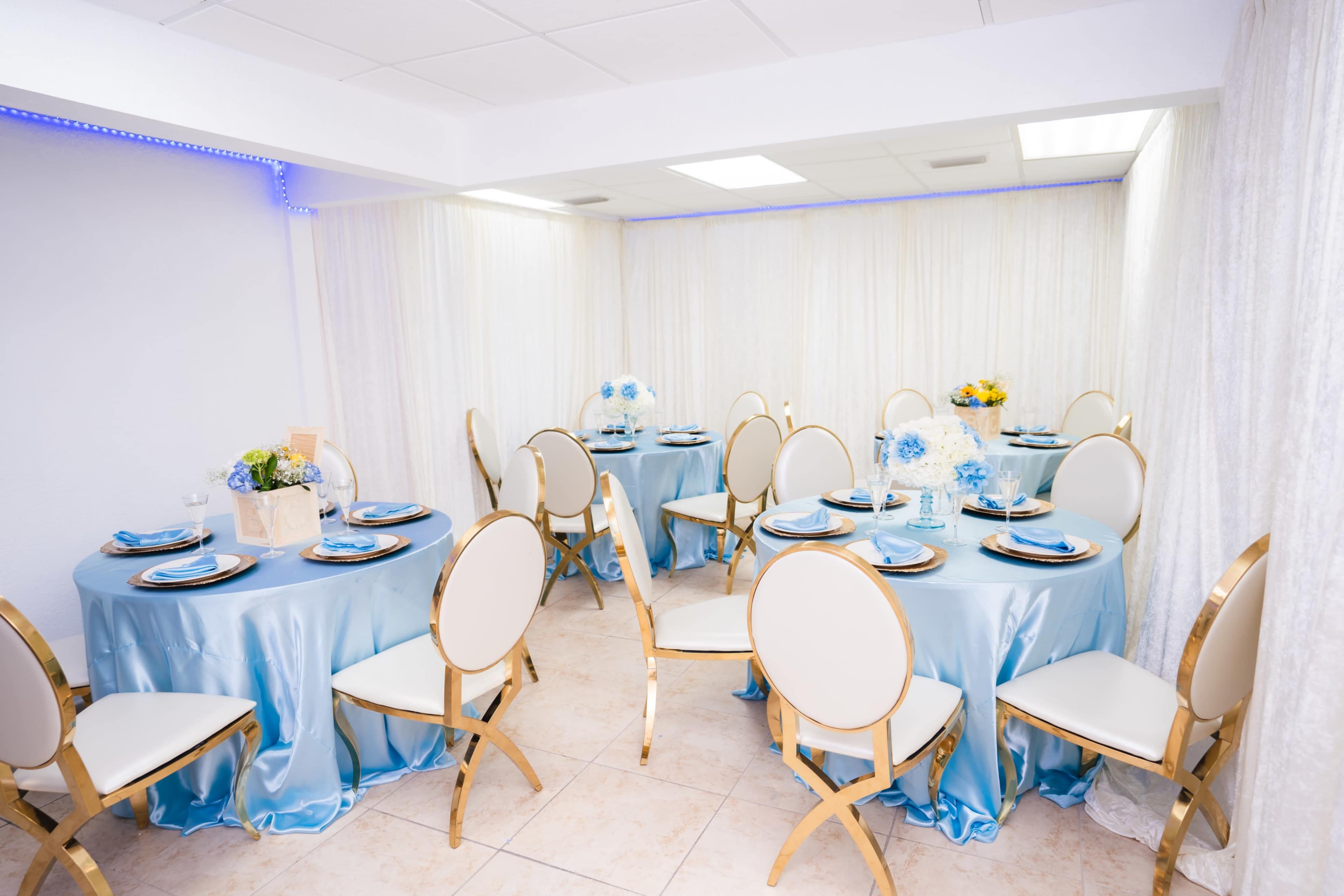 The image shows a brightly lit banquet room set up with round tables covered in light blue tablecloths, surrounded by gold-accented chairs.