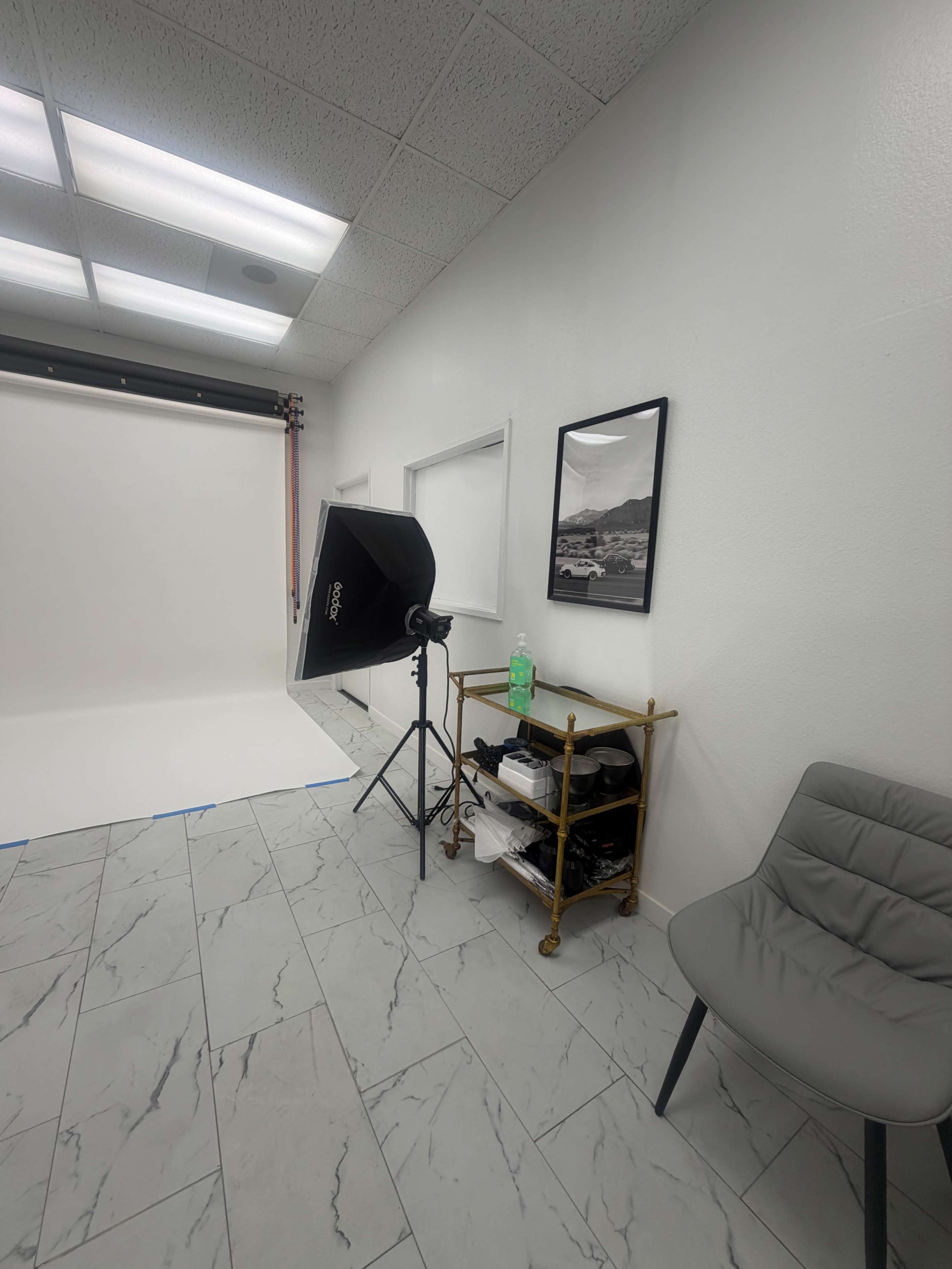 The image shows a well-lit photography studio featuring a backdrop setup, a light source on a stand, and a small table with various items in a corner.
