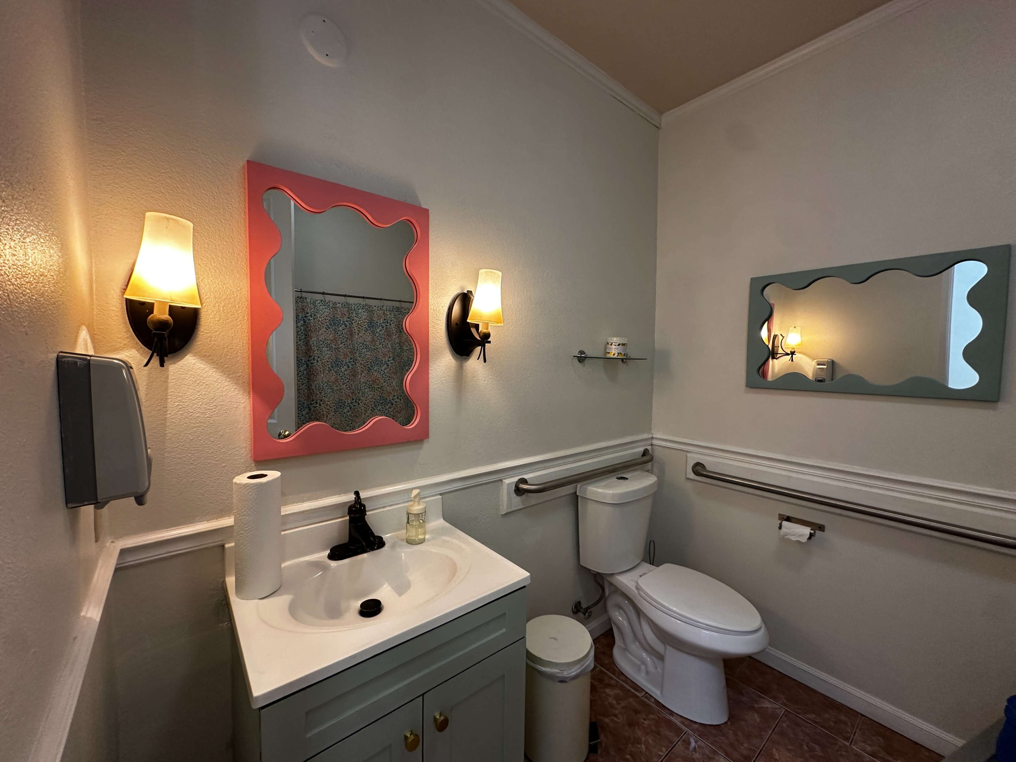 The image shows a small bathroom featuring a sink with a light green cabinet, two mirrors with decorative frames, a toilet, and wall-mounted light fixtures.