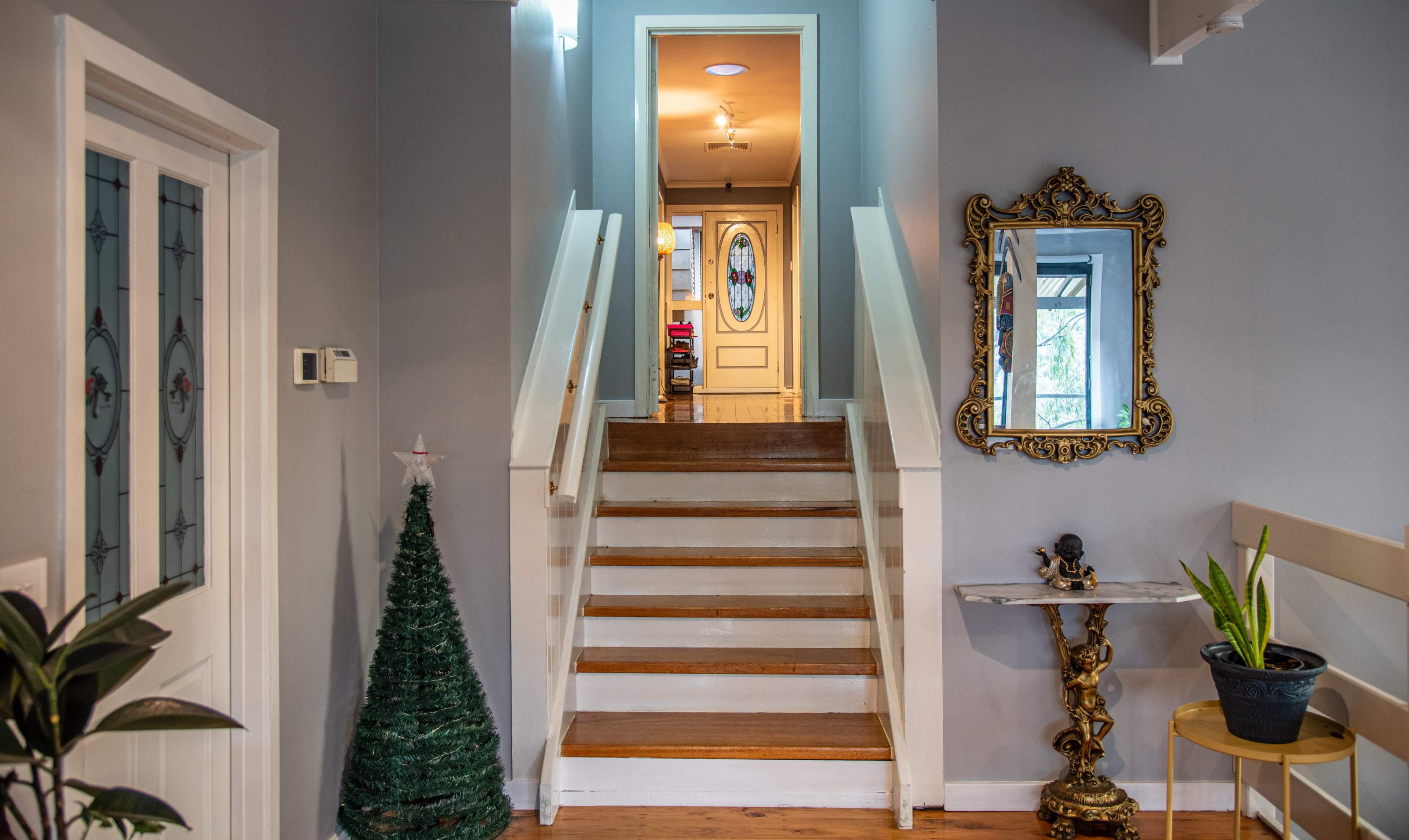 The image shows a staircase leading to an upper level, framed by a decorative mirror and a small Christmas tree in a brightly lit hallway.