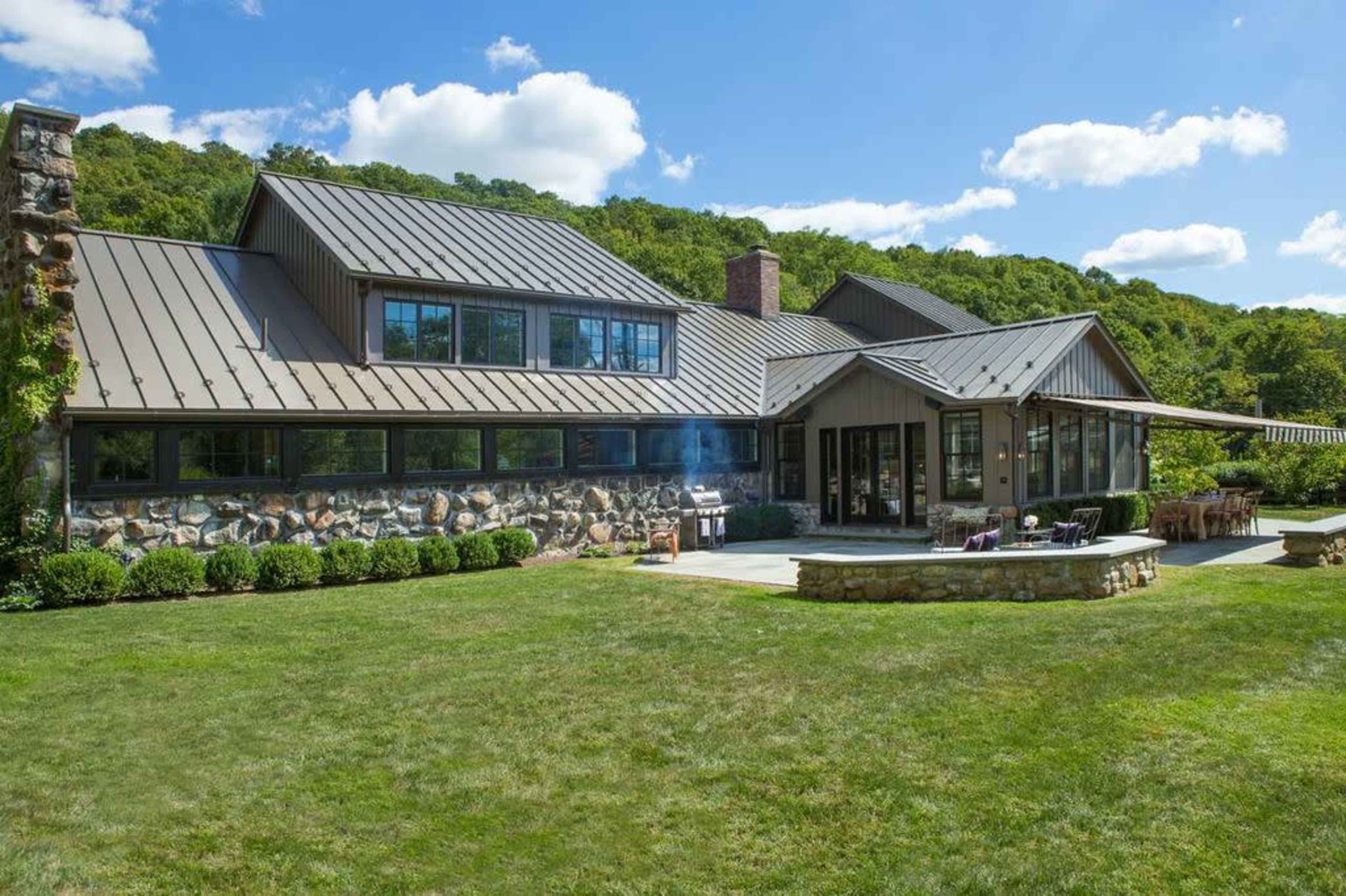 A modern, multi-level house with a stone and metal exterior is situated on a grassy landscape surrounded by trees.