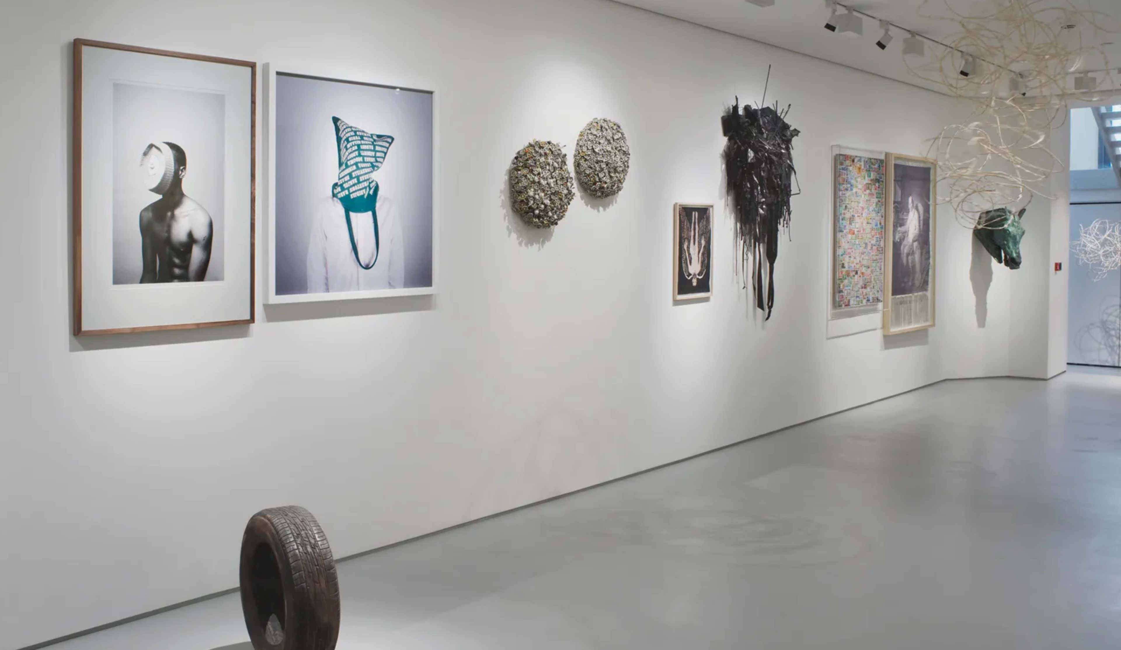 The image shows a contemporary art gallery with a white wall displaying various framed artworks and sculptures.