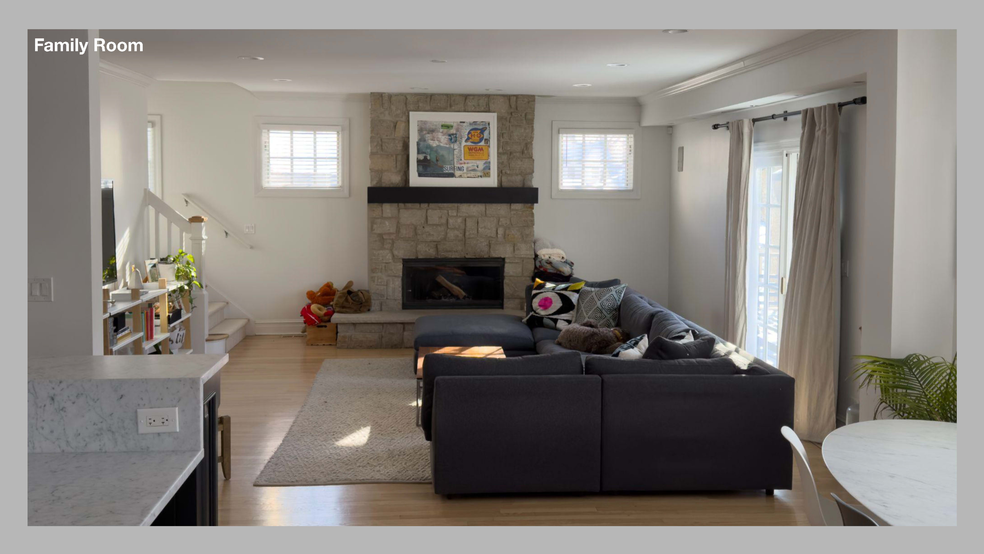 The image shows a family room featuring a large sectional sofa, a stone fireplace, and sunlight streaming in through the windows.
