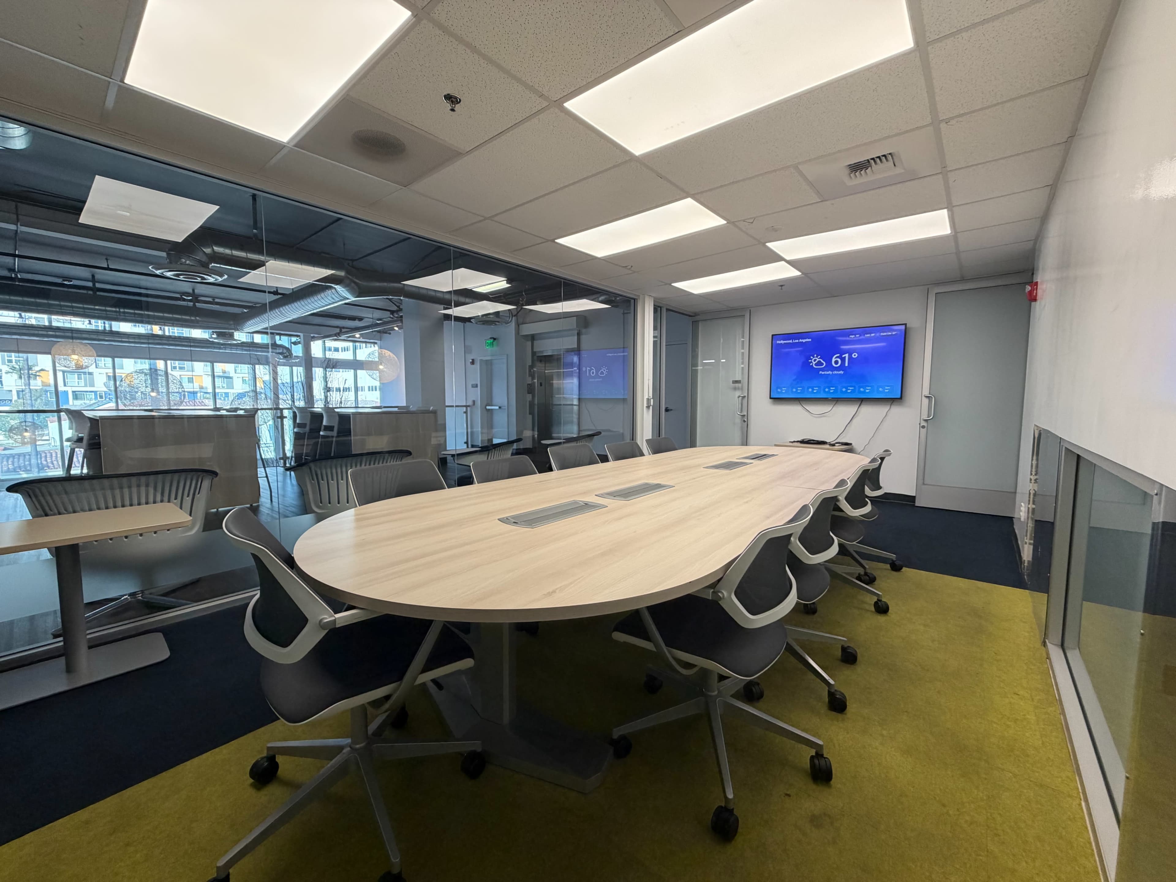 The image shows a modern conference room with a large oval table, ergonomic chairs, and a display screen mounted on the wall.