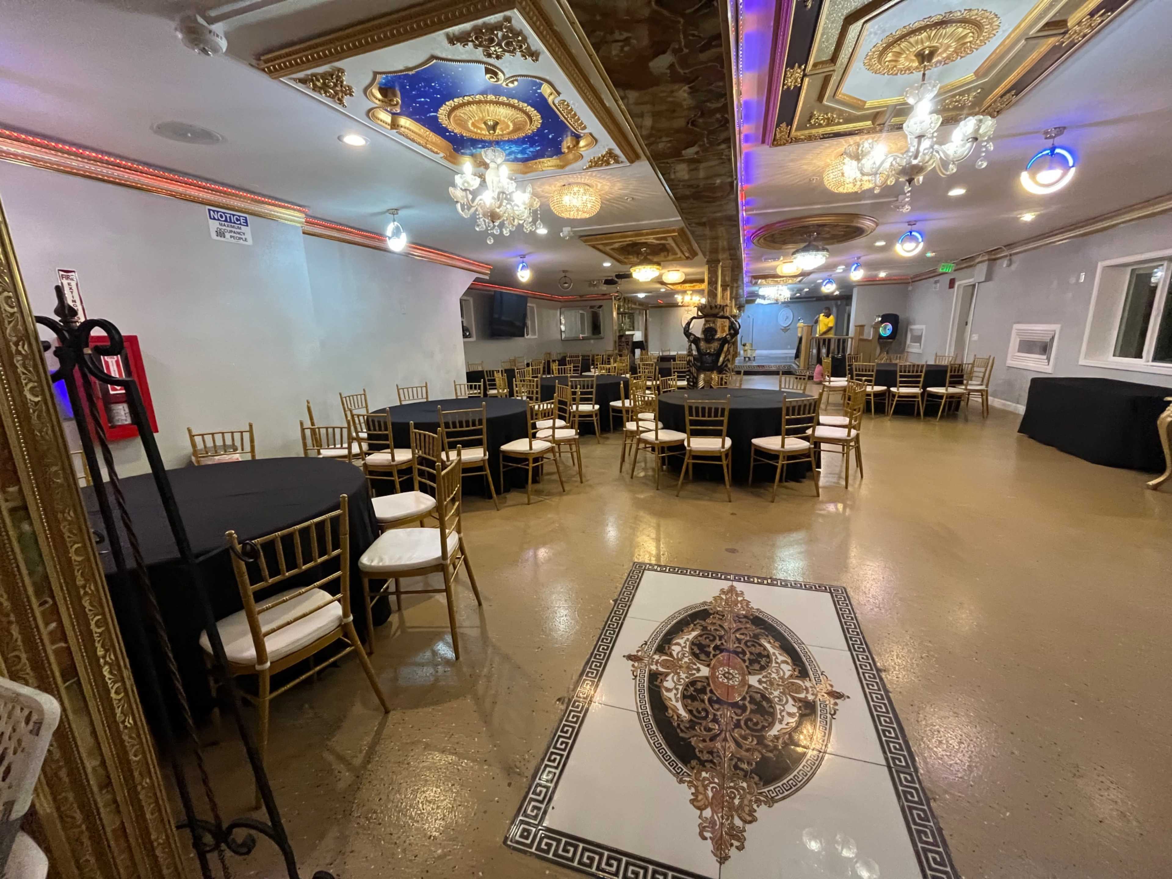 The image shows a banquet hall with several round tables covered in black tablecloths, decorated with gold chairs, and featuring ornate ceiling designs and chandeliers.