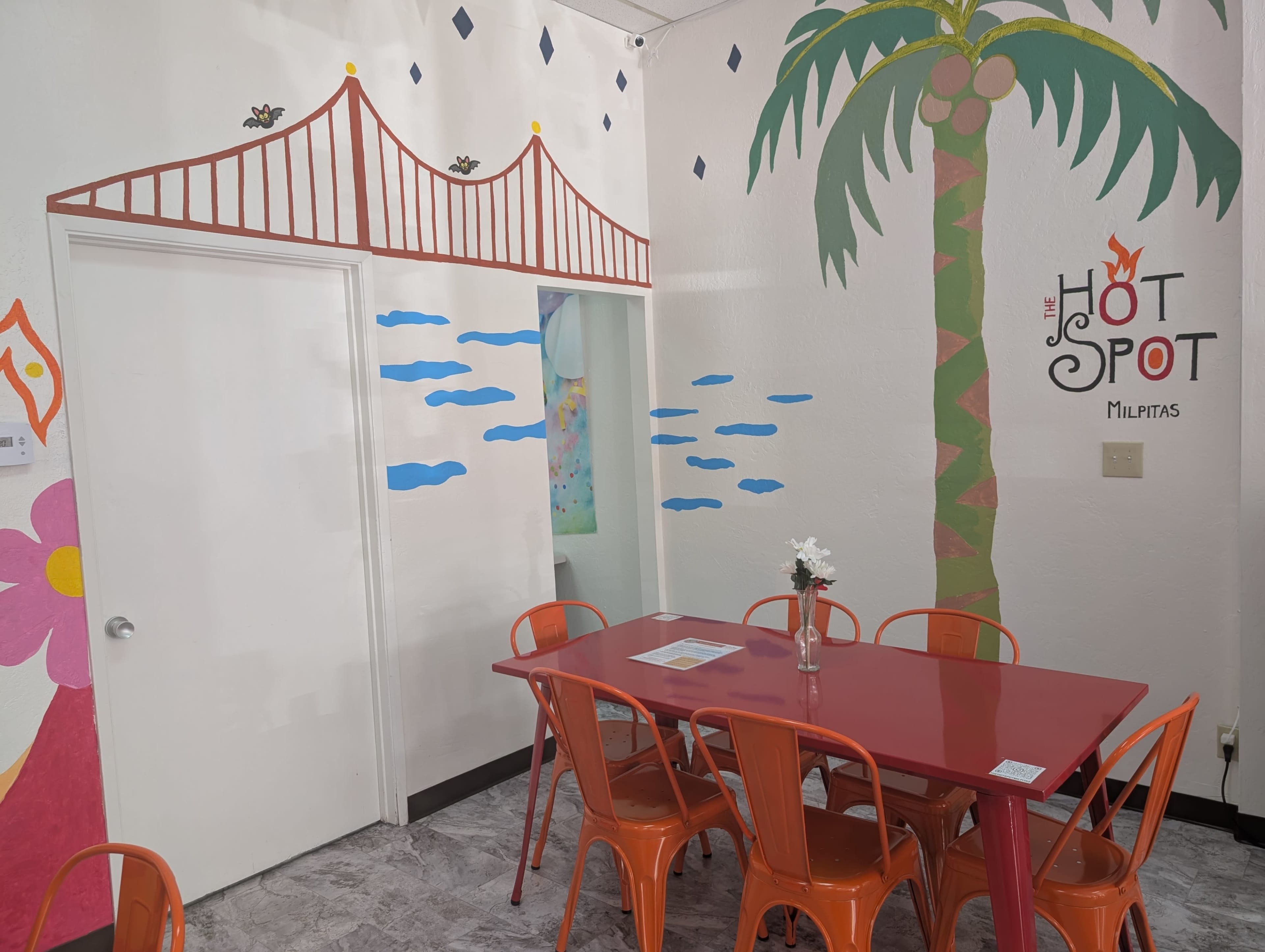 The image shows a brightly decorated dining area with a red table and orange chairs, featuring murals of a palm tree and geometric designs on the walls.