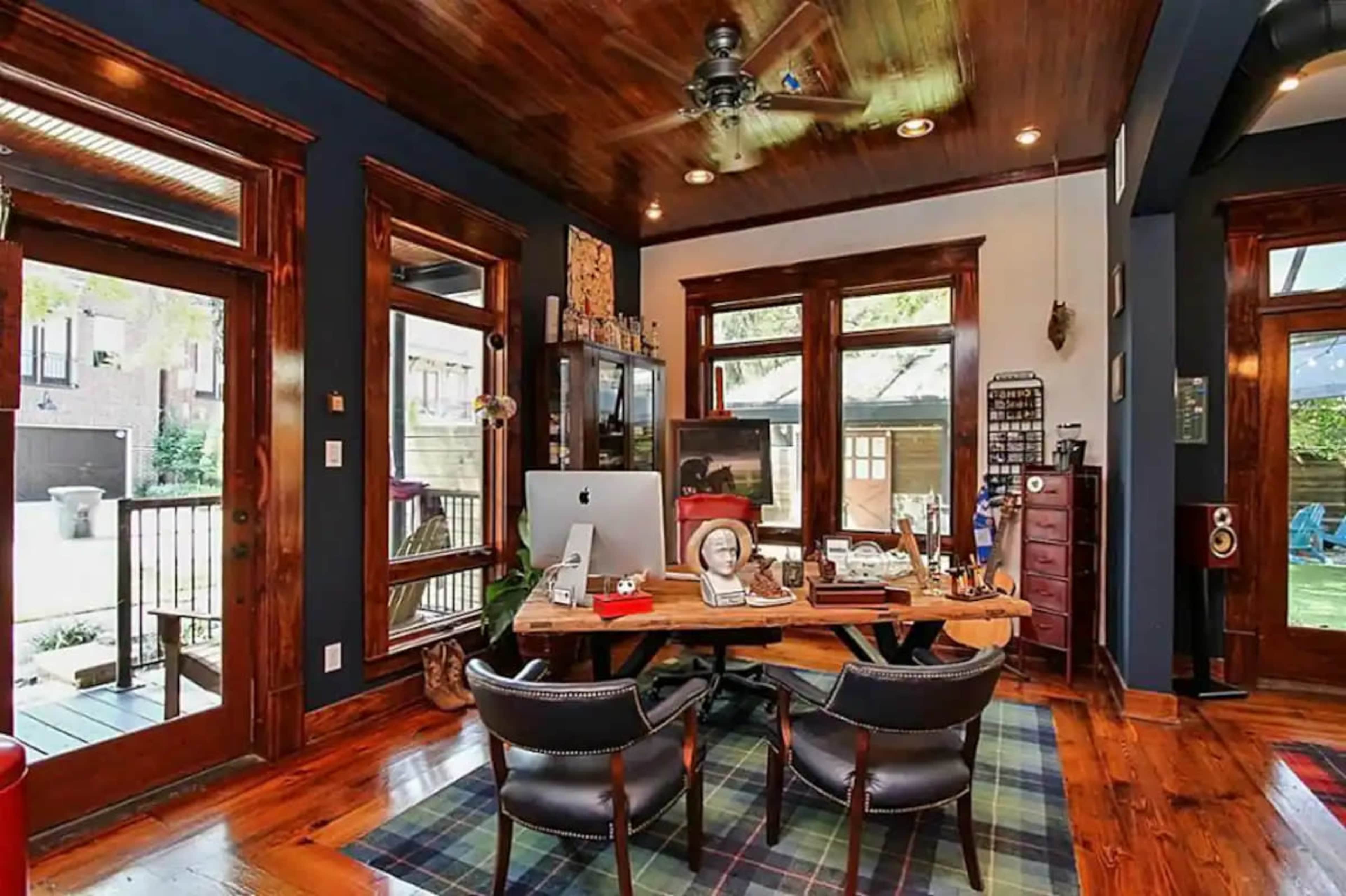 A wooden home office with a large desk facing a computer, flanked by two chairs, and surrounded by windows that provide natural light.