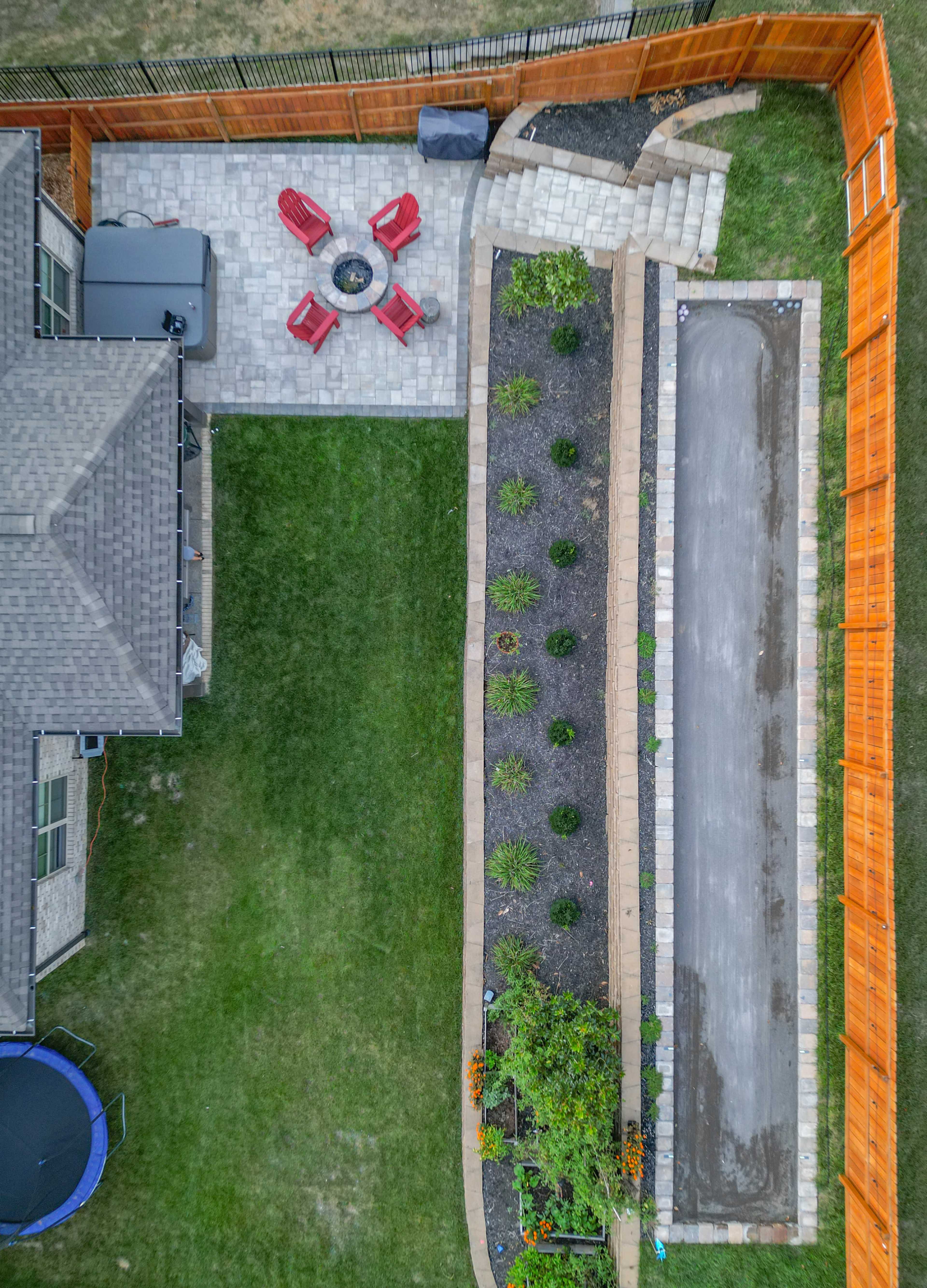 An aerial view shows a backyard with a paved seating area featuring four red chairs around a fire pit, a garden along one side, and a gravel pathway leading to a staircase.