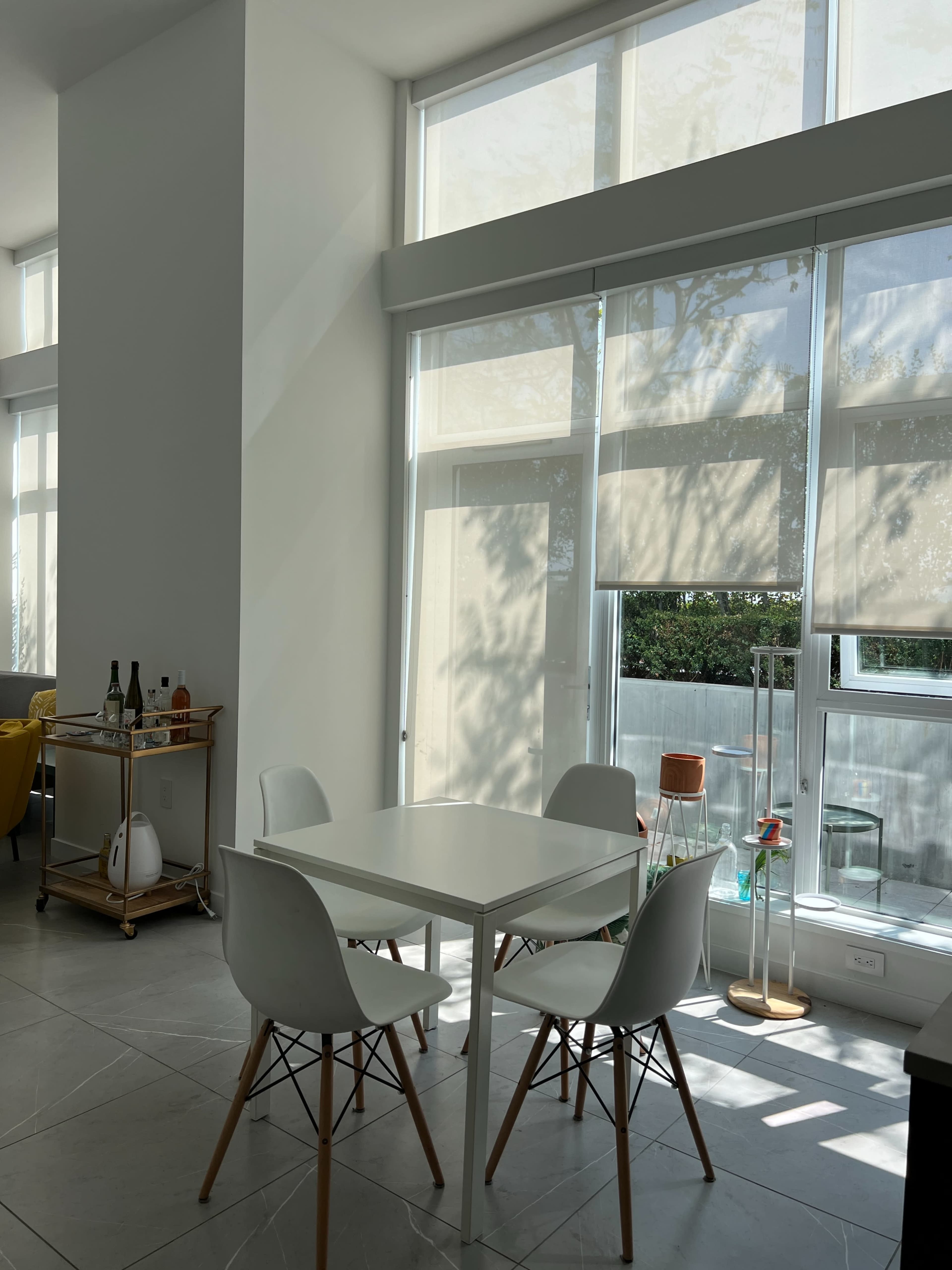 The image shows a modern dining area with a white table surrounded by four chairs, large windows allowing natural light, and a cart with wine bottles in the background.