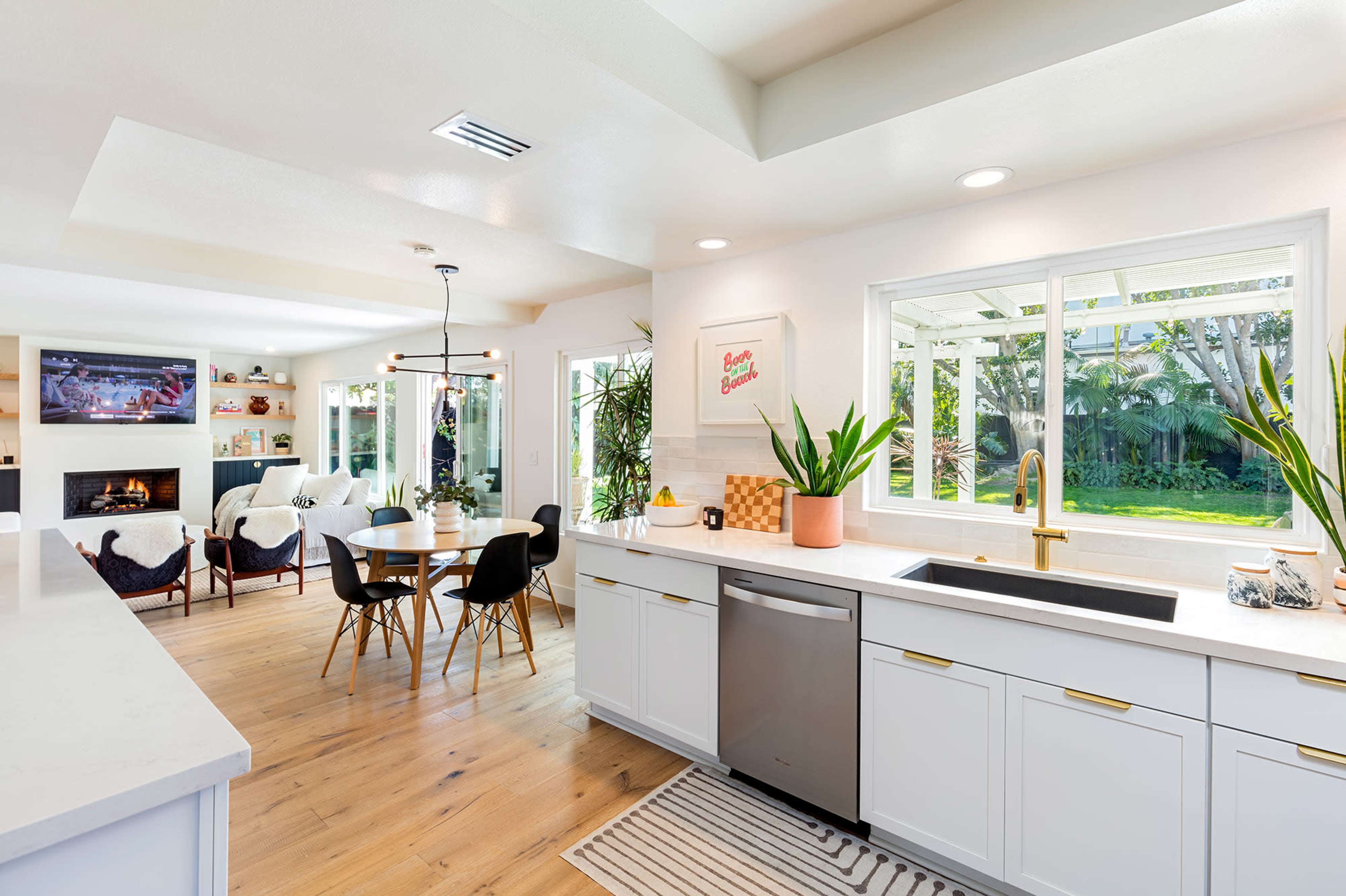 A modern kitchen with white cabinets and a sink, offering a view into a bright living area featuring a television and seating.