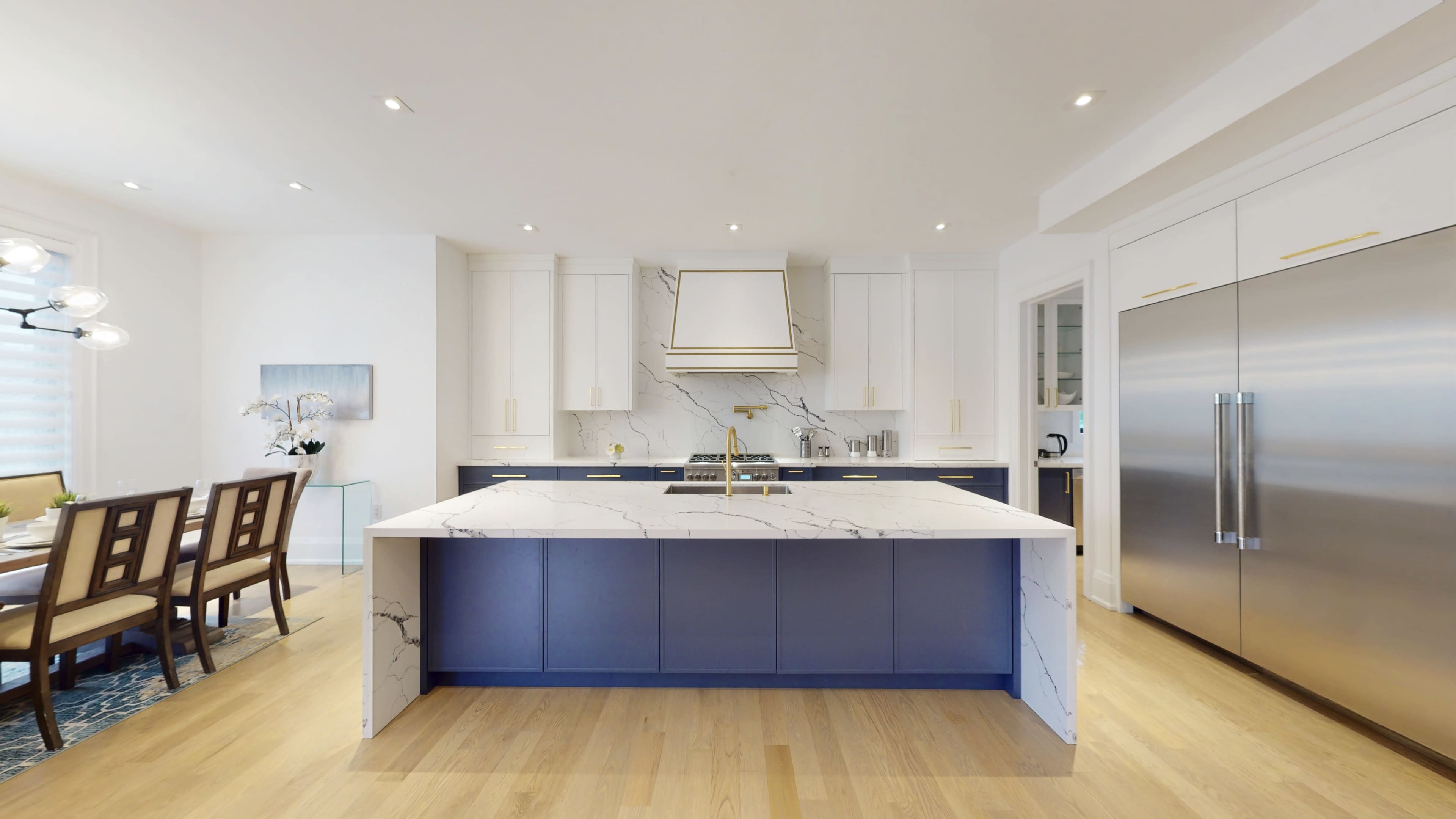 A modern kitchen features a large marble island with a blue base, white cabinetry, and stainless steel appliances.