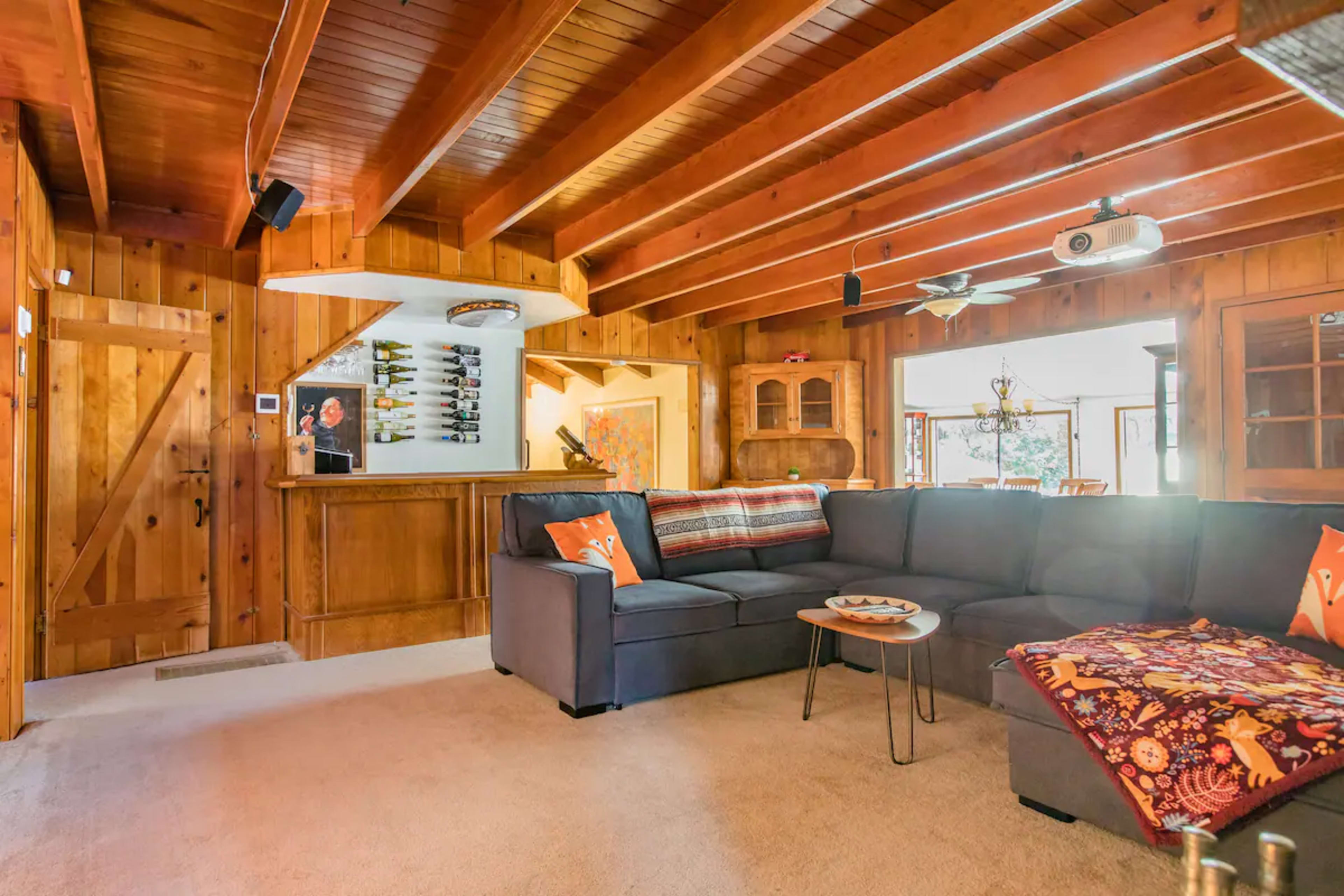 The image shows a cozy living room with wooden walls, a sectional sofa, a small table, and a bar area in the background.