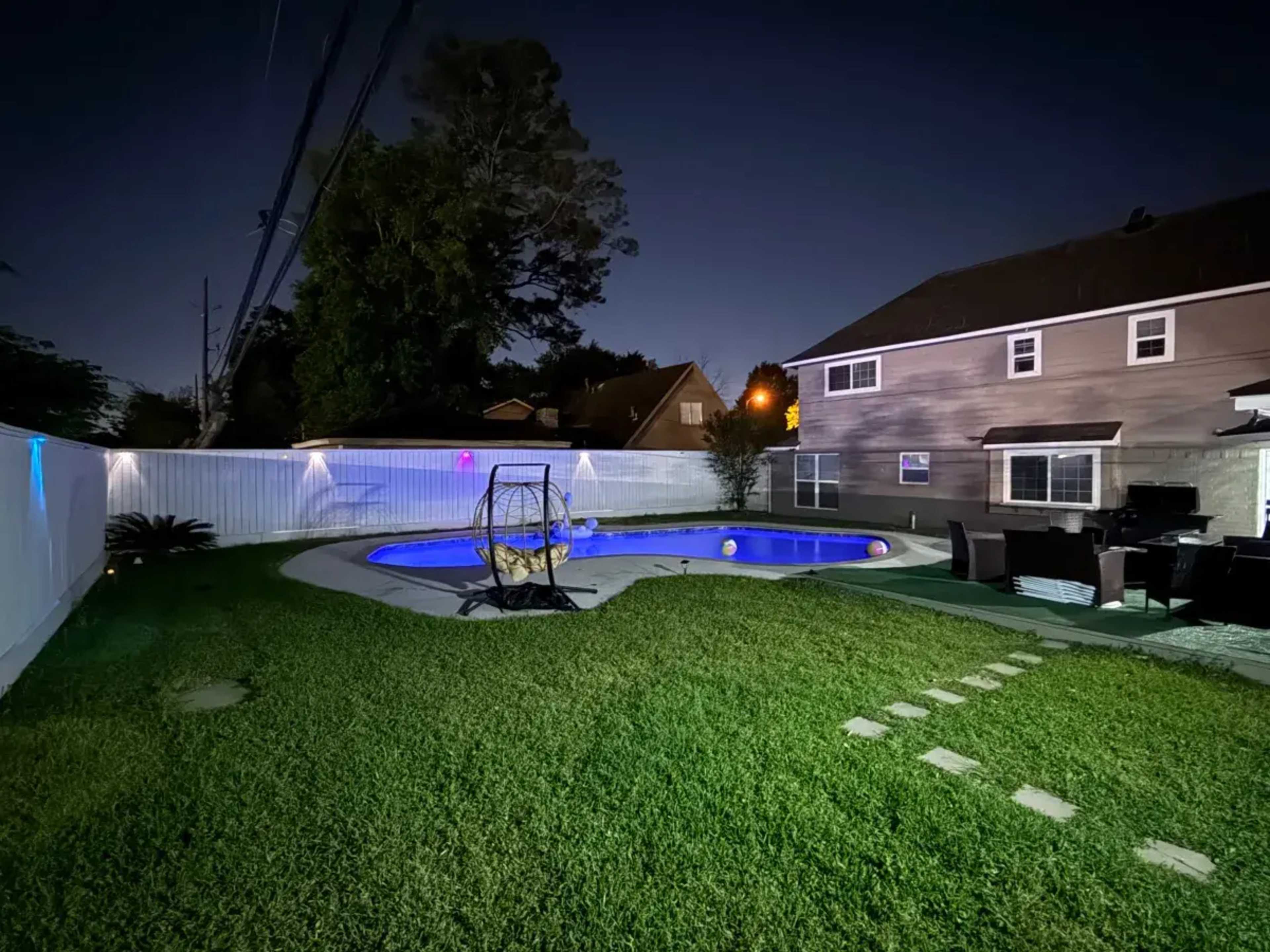 The image shows a backyard at night with a swimming pool, a grassy area, a swing set, and outdoor seating near a house.