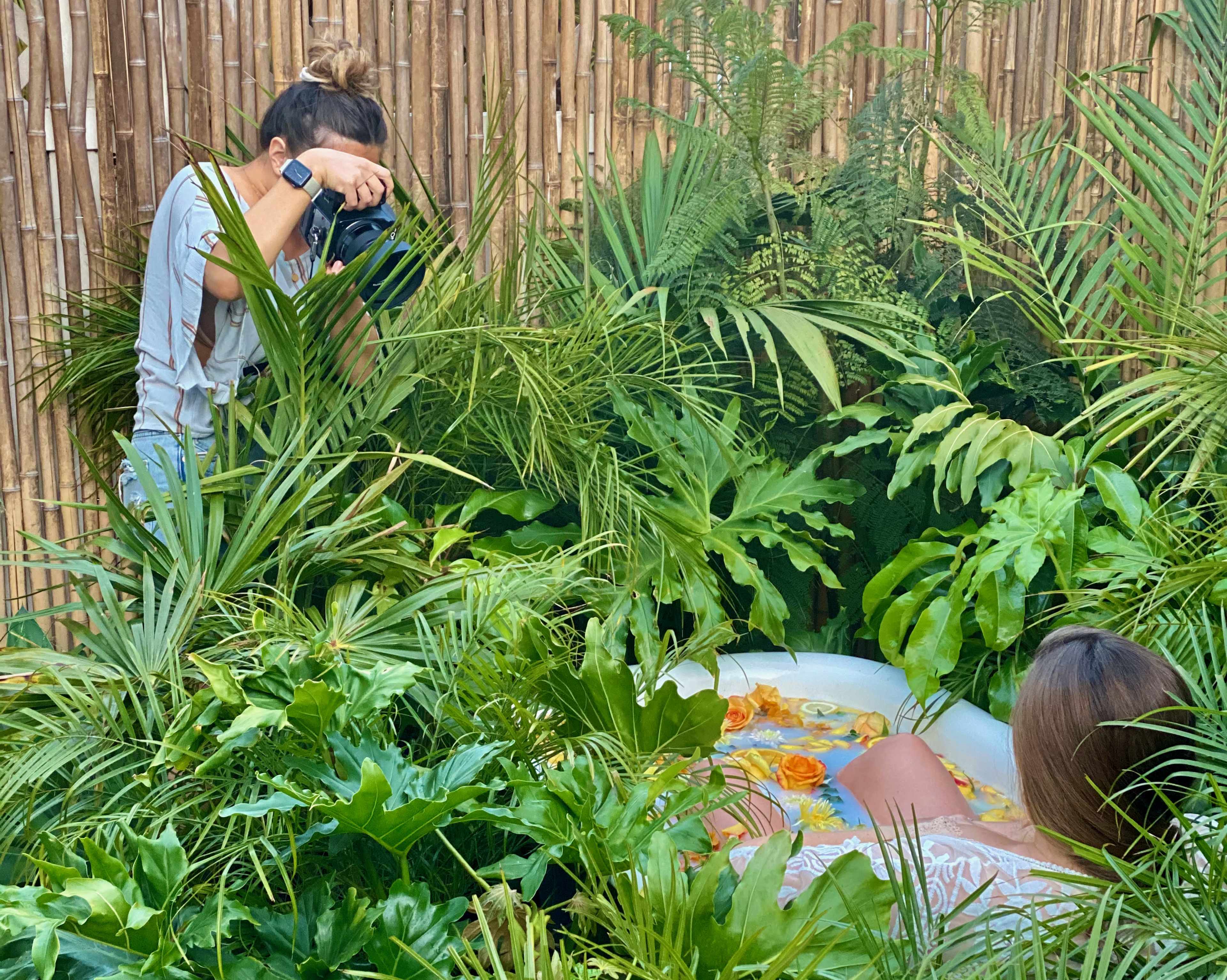 A woman is taking photos of another woman relaxing in a bathing tub surrounded by lush greenery.