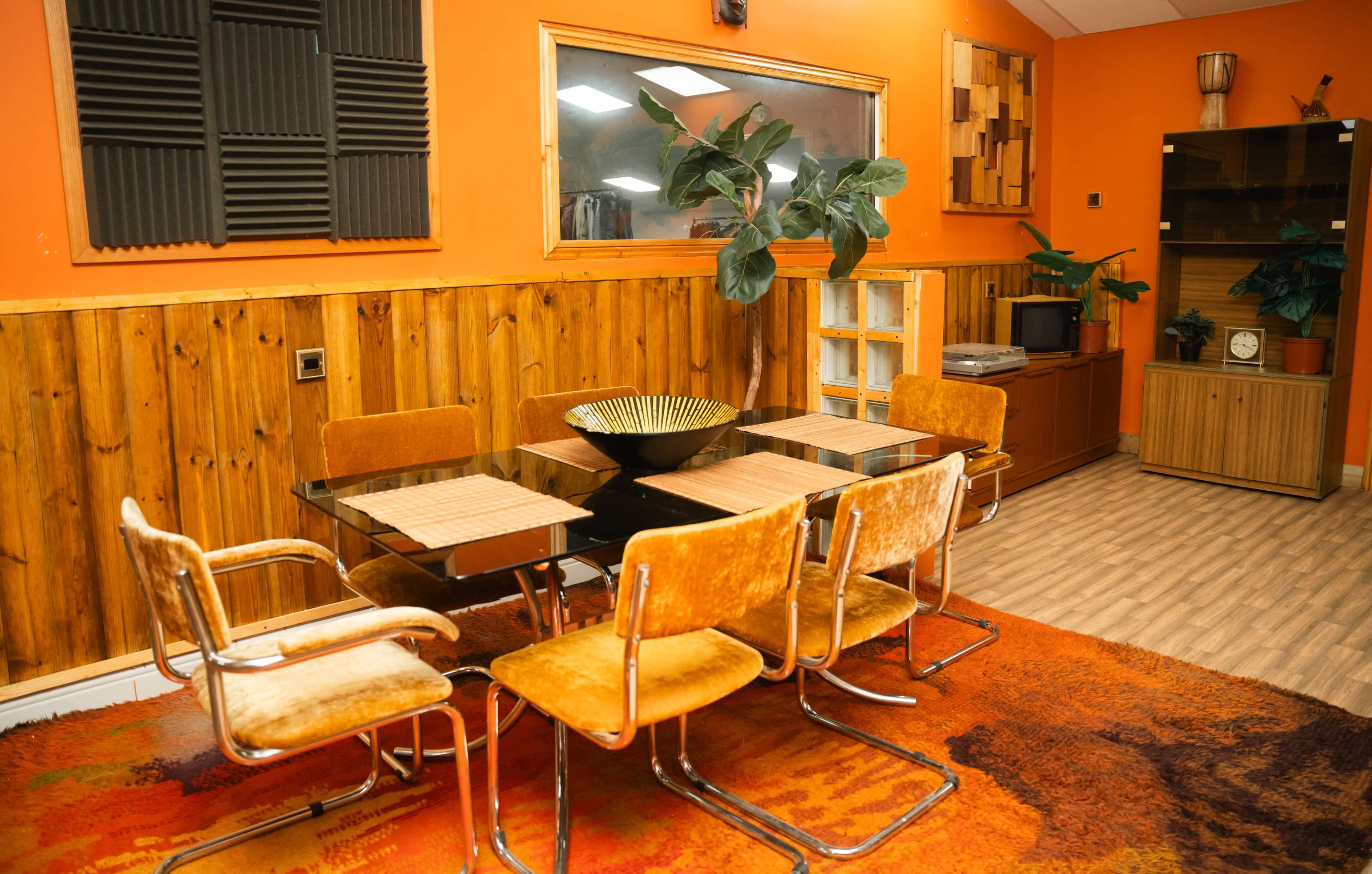 A modern dining area with a black glass table surrounded by six orange-brown chairs, set against a backdrop of wood paneling and an orange wall, featuring plants and decorative shelves.