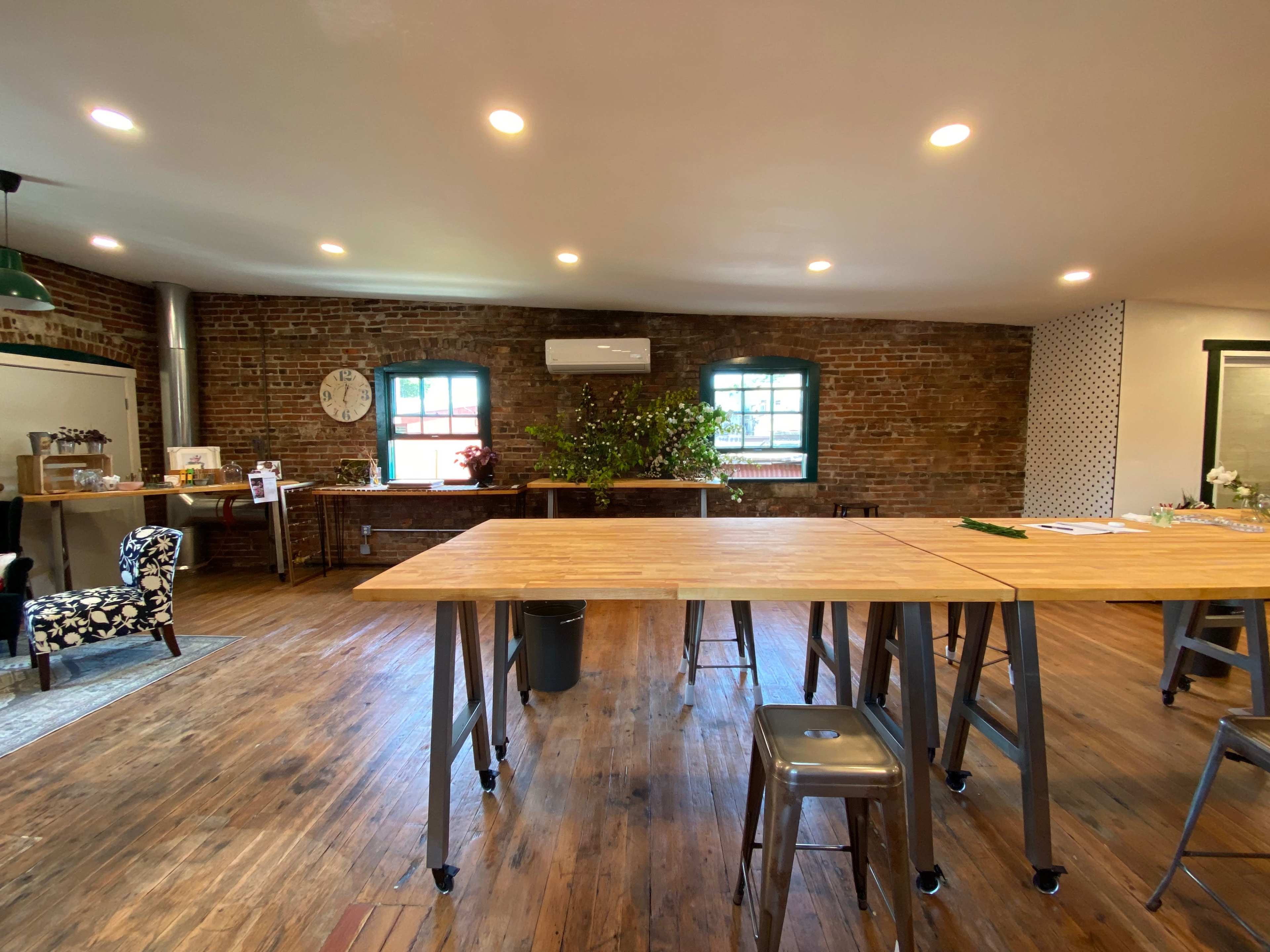 A well-lit, spacious room with wooden floors and exposed brick walls, featuring several large tables and a small greenery arrangement in the corner.