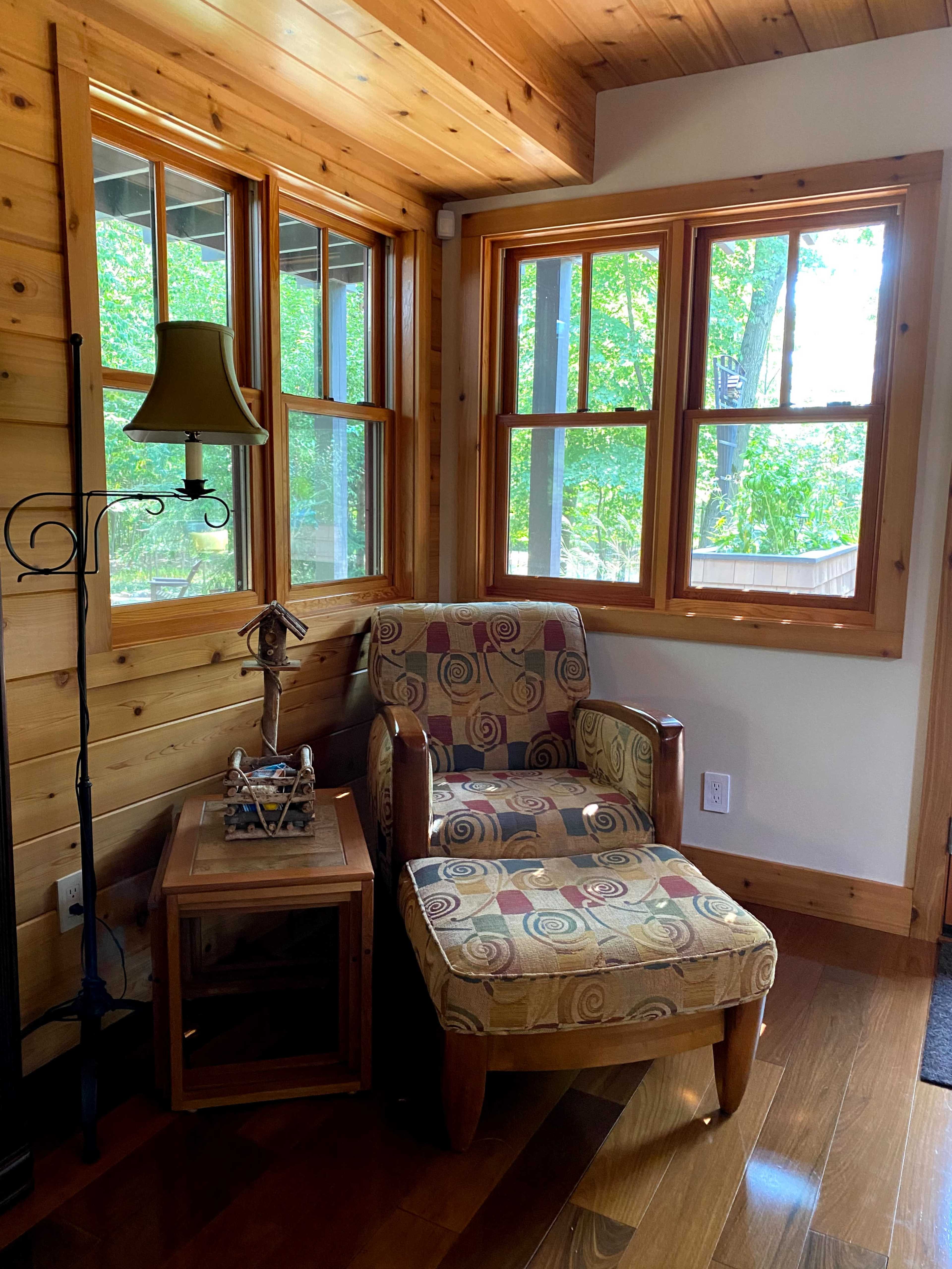 A cozy corner features a patterned armchair beside a wooden side table and a lamp, with windows allowing natural light to enter.