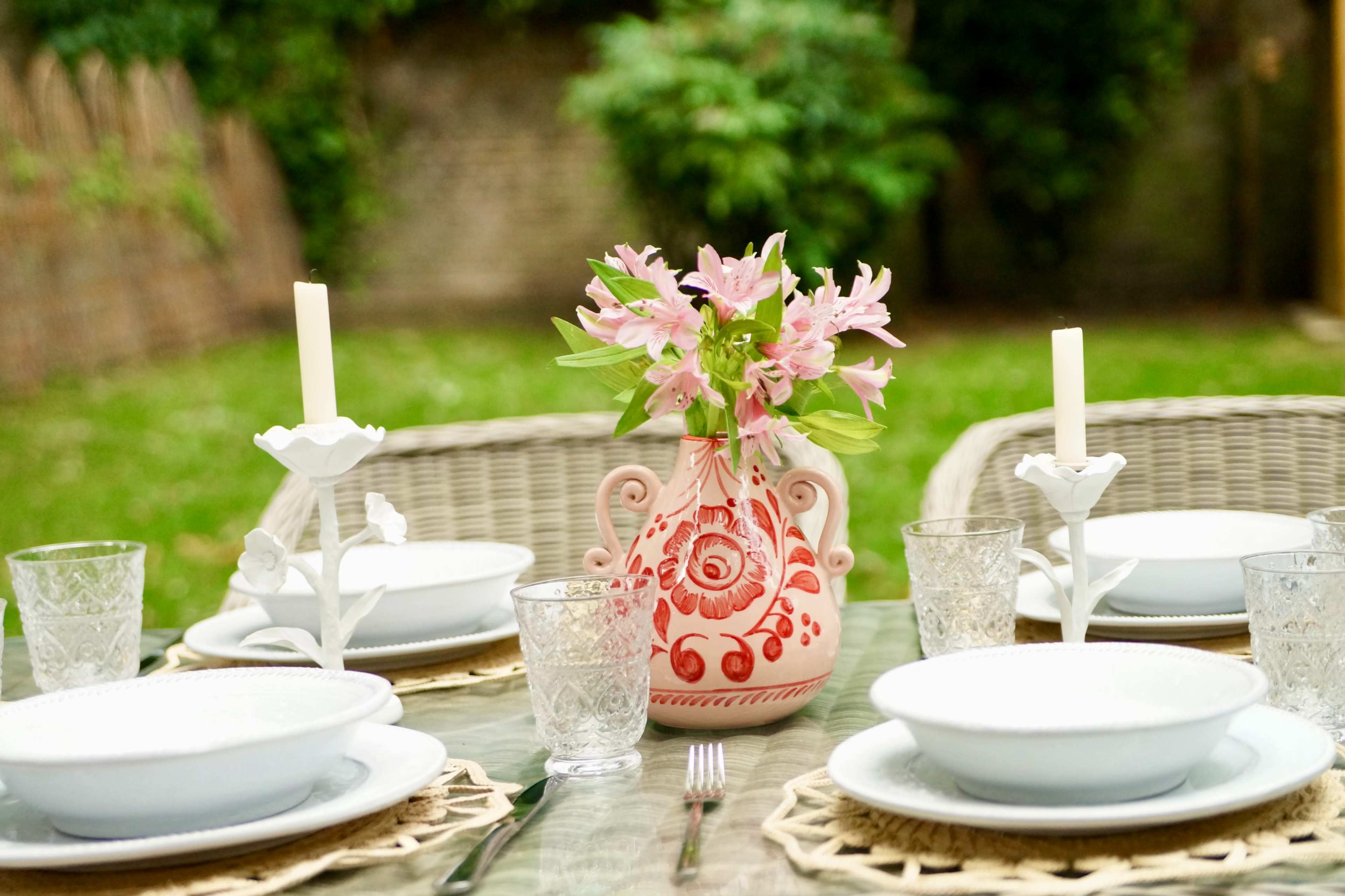 A well-set outdoor dining table features white plates, elegant glassware, and a decorative vase filled with pink flowers at its center.