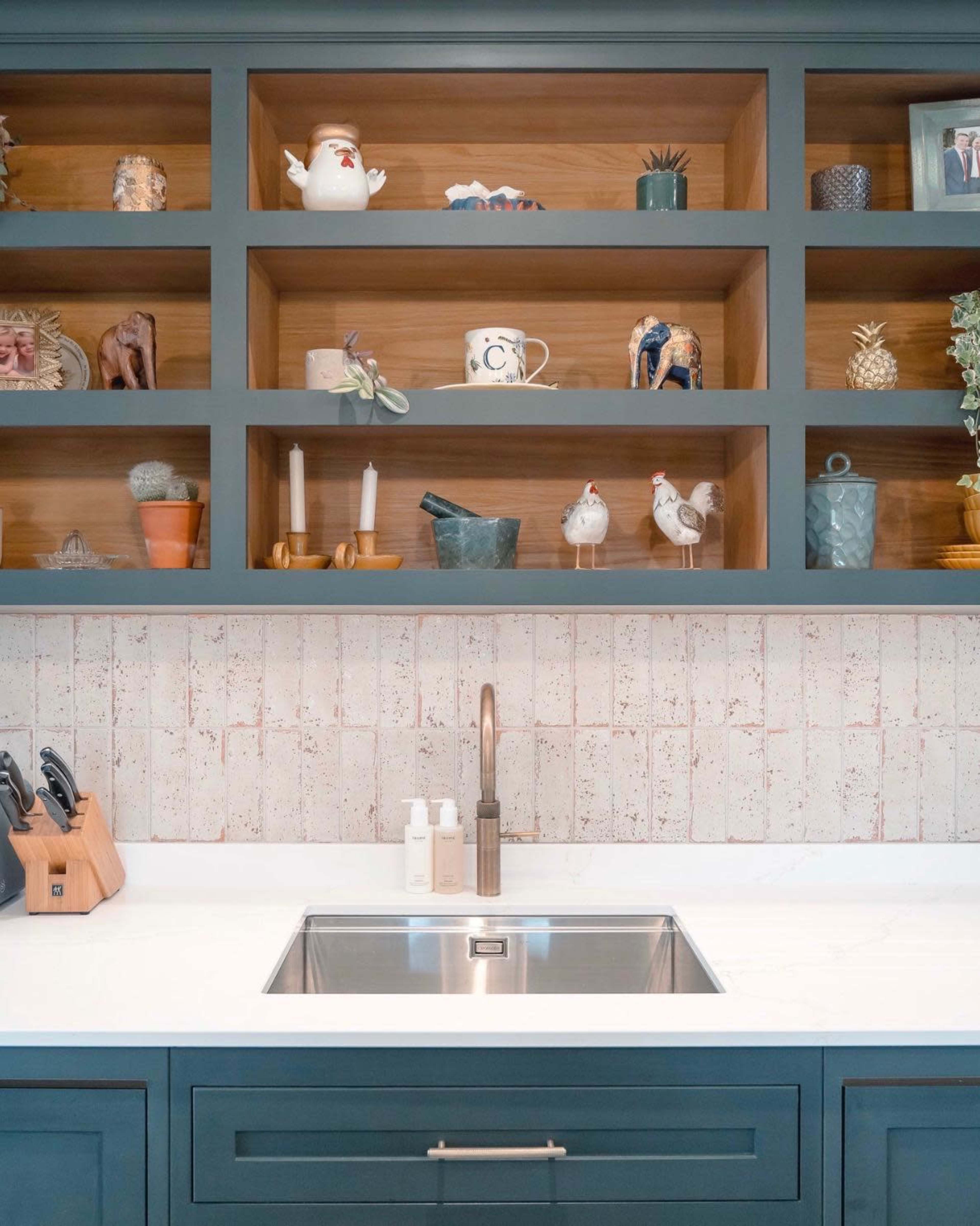 The image shows a kitchen with open shelving displaying various decorative items and a sink with a modern faucet beneath a white countertop.