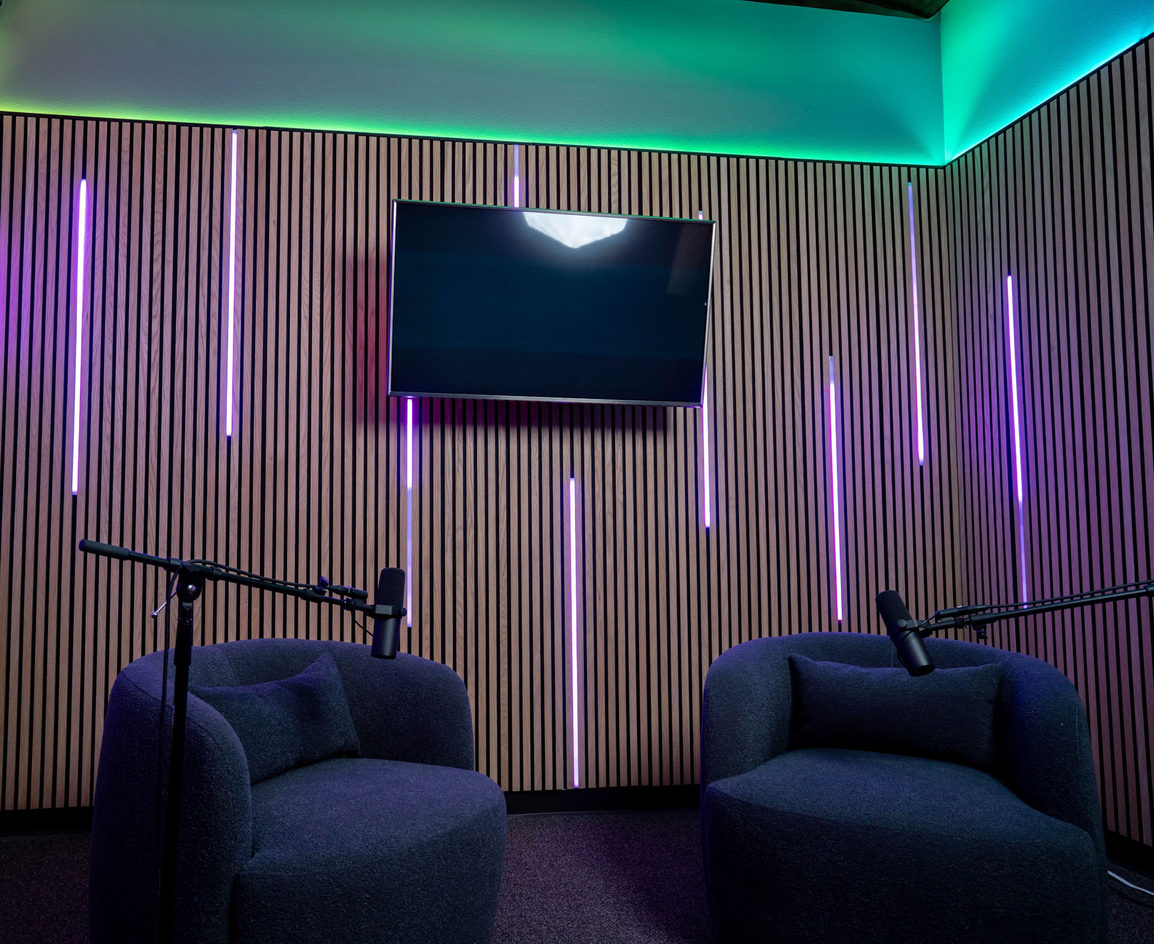 A soundproofed recording room features two rounded chairs, each with a microphone, and a wall adorned with vertical light strips and a mounted television.