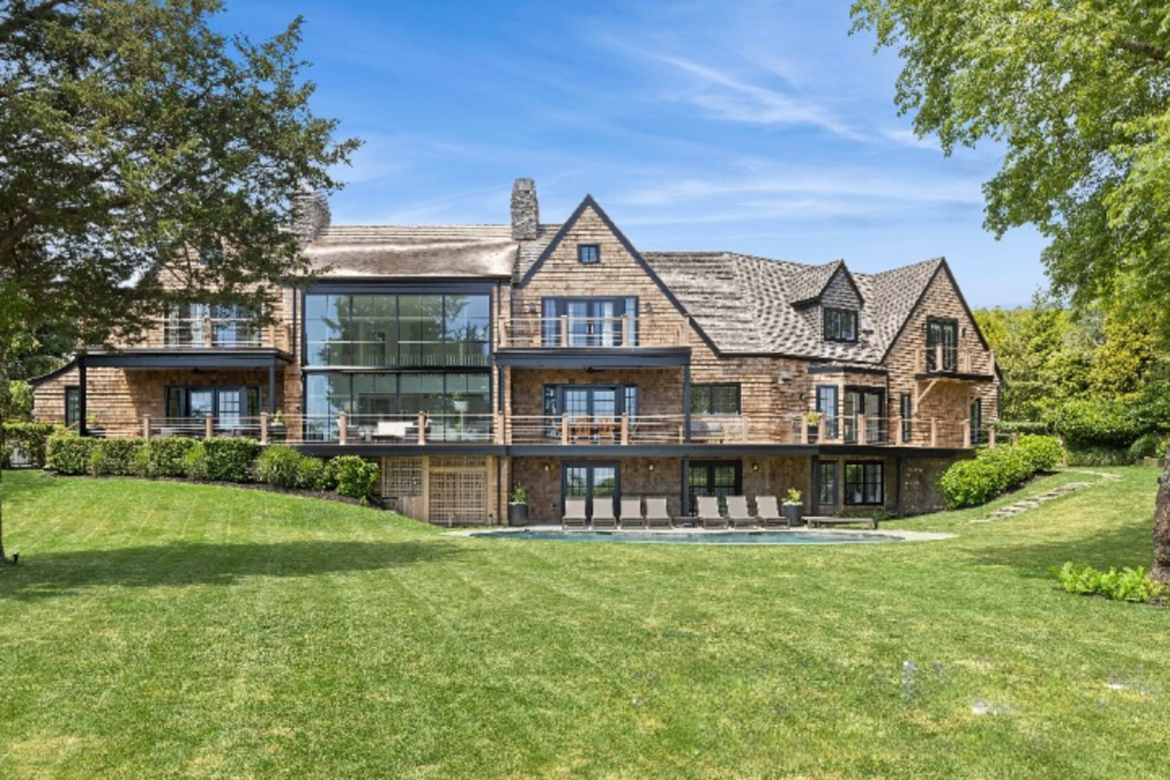 A large wooden house with multiple balconies set on a grassy hillside, surrounded by trees and a clear blue sky.