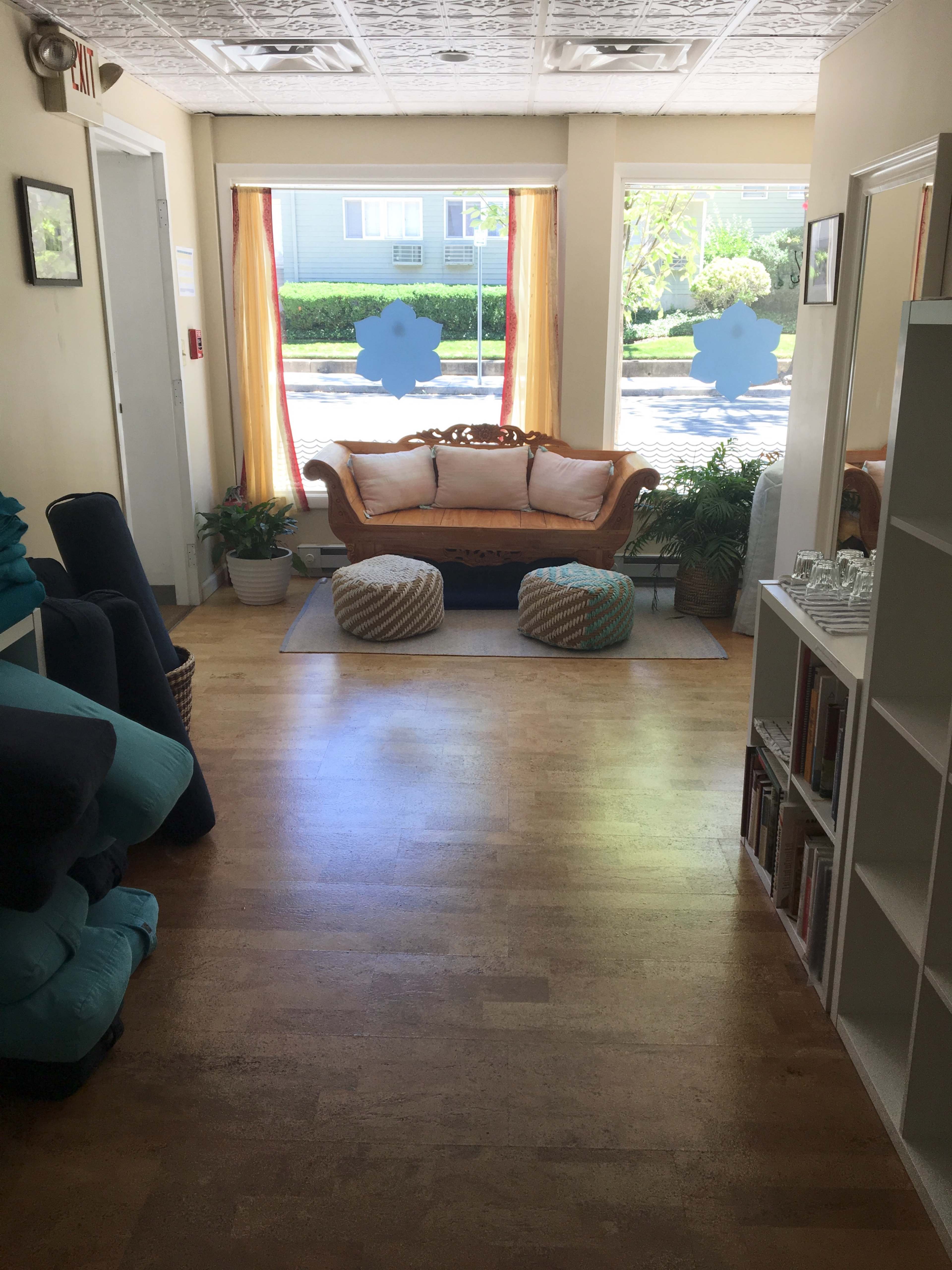 A light-filled interior of a welcoming space with a brown couch, two round poufs, and potted plants near large windows.