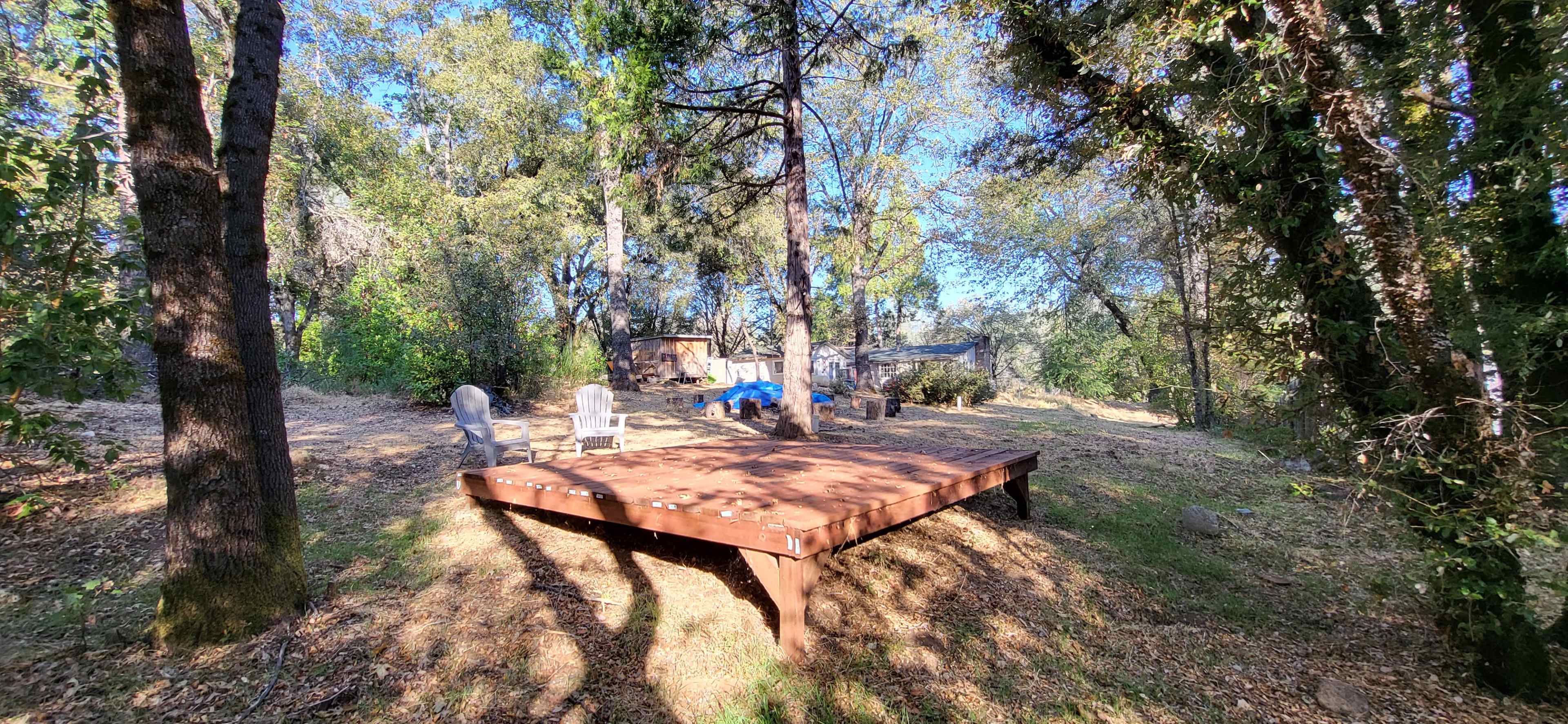 The image shows a wooden platform surrounded by trees, with several chairs positioned nearby in a natural setting.