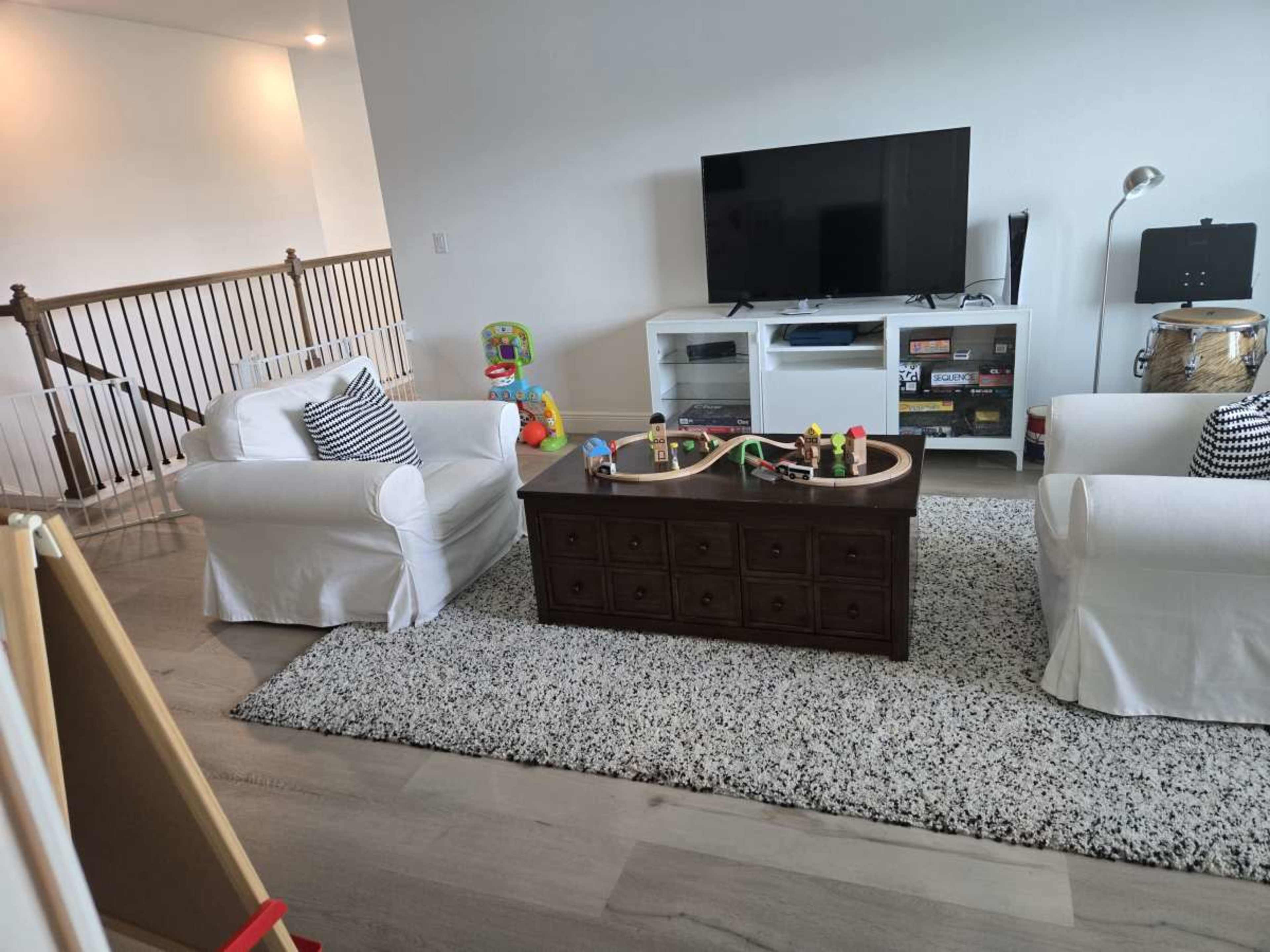 The image shows a living room with two white armchairs, a dark wooden coffee table displaying toys and a train set, a television on a stand, and an easel in the corner.
