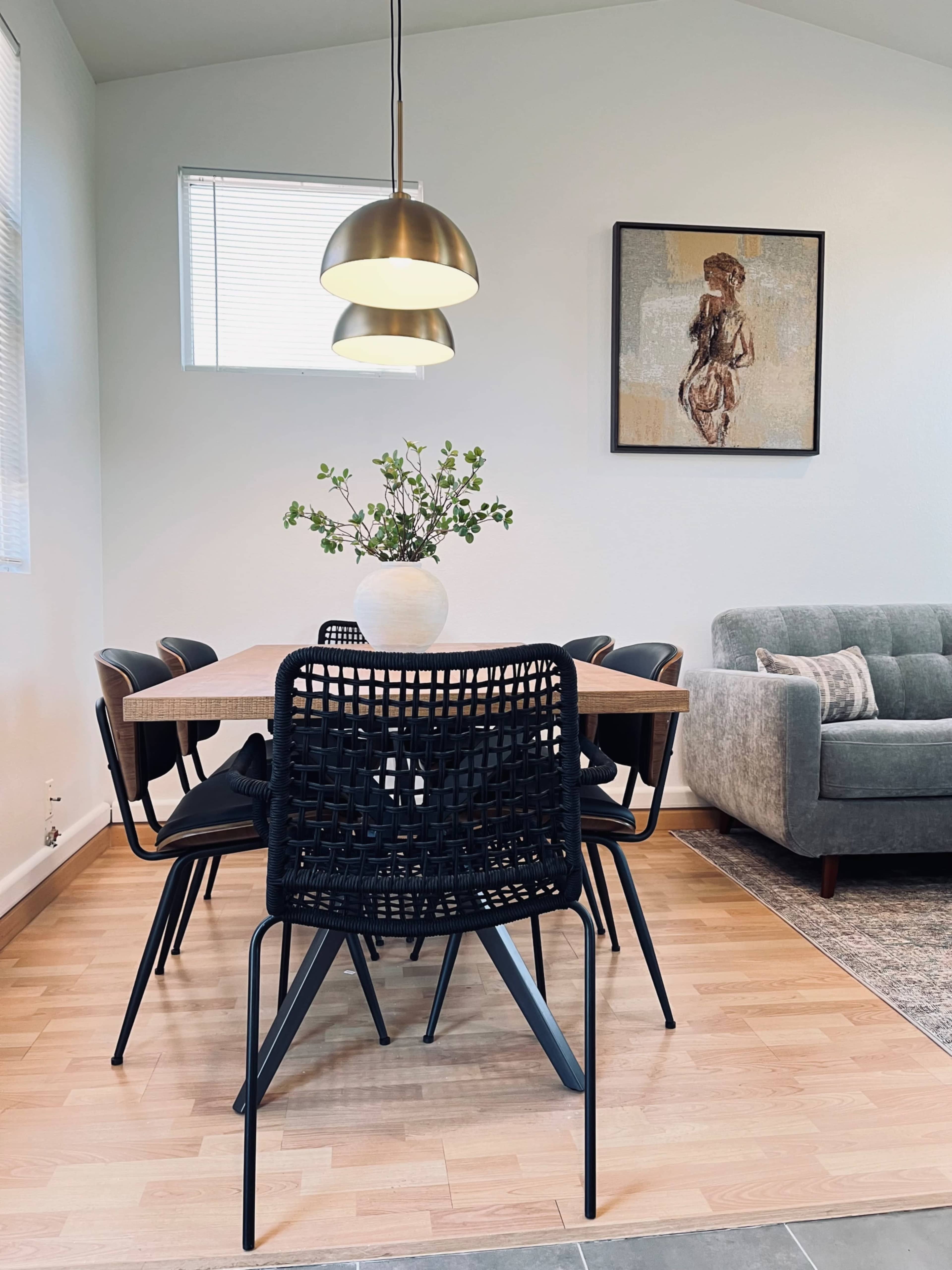 A dining area features a wooden table surrounded by black chairs, a decorative vase with greenery, and a sofa in the corner.