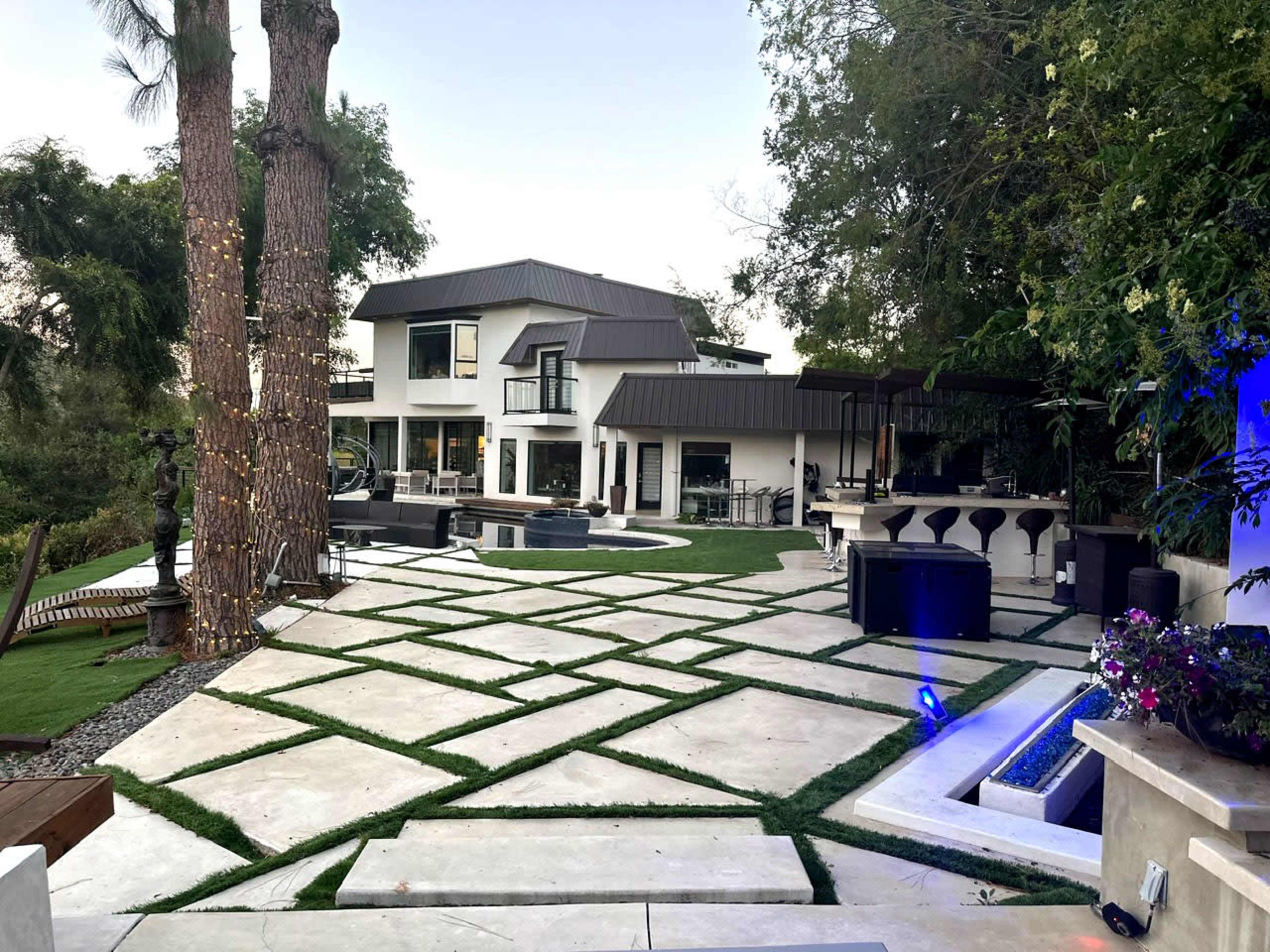 A modern two-story house surrounded by a landscaped yard with geometric stone pathways and patio areas featuring outdoor seating and a barbecue setup.