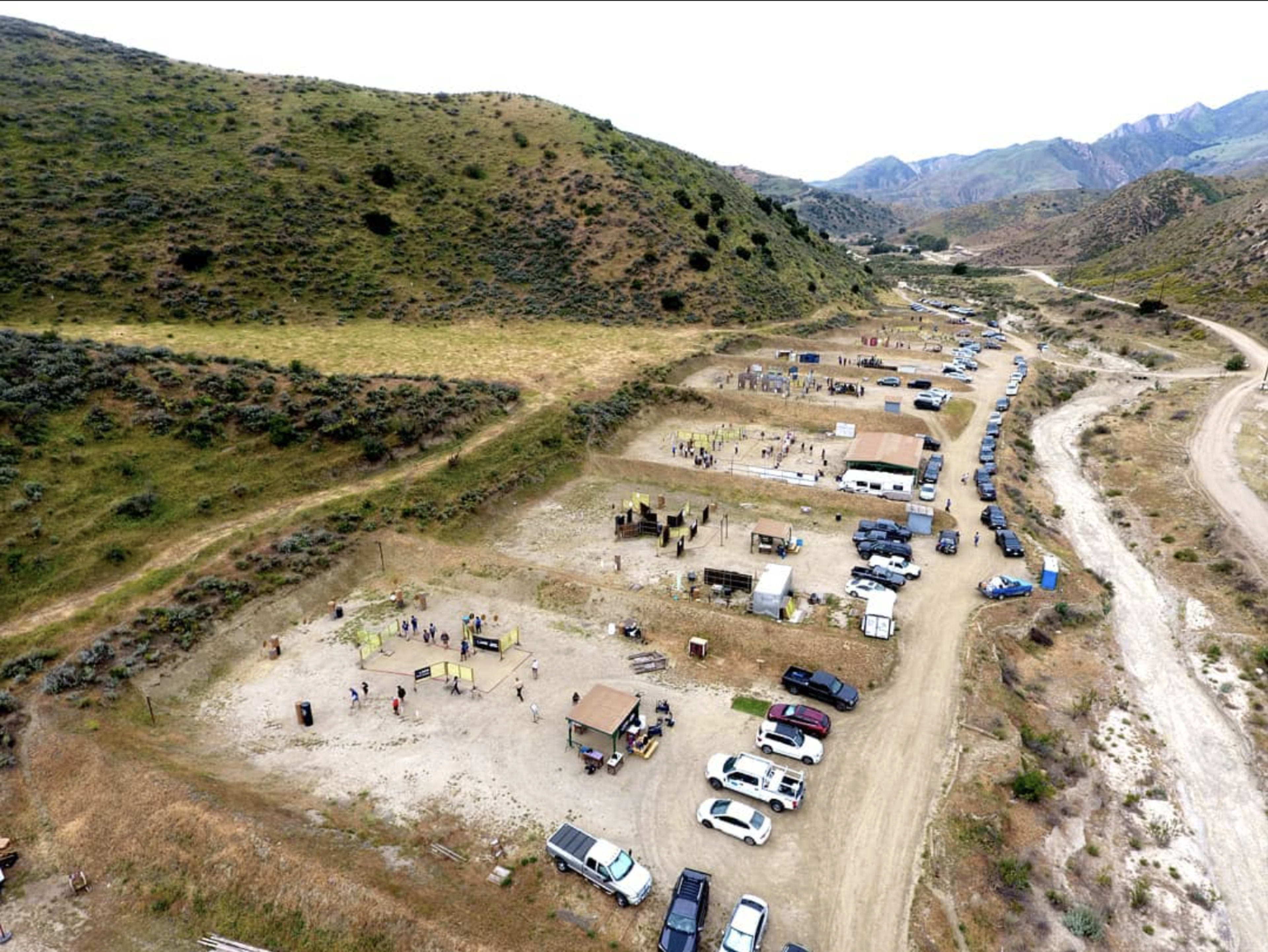 The image shows an outdoor activity area with several wooden structures, vehicles parked along a dirt road, and a hilly landscape in the background.