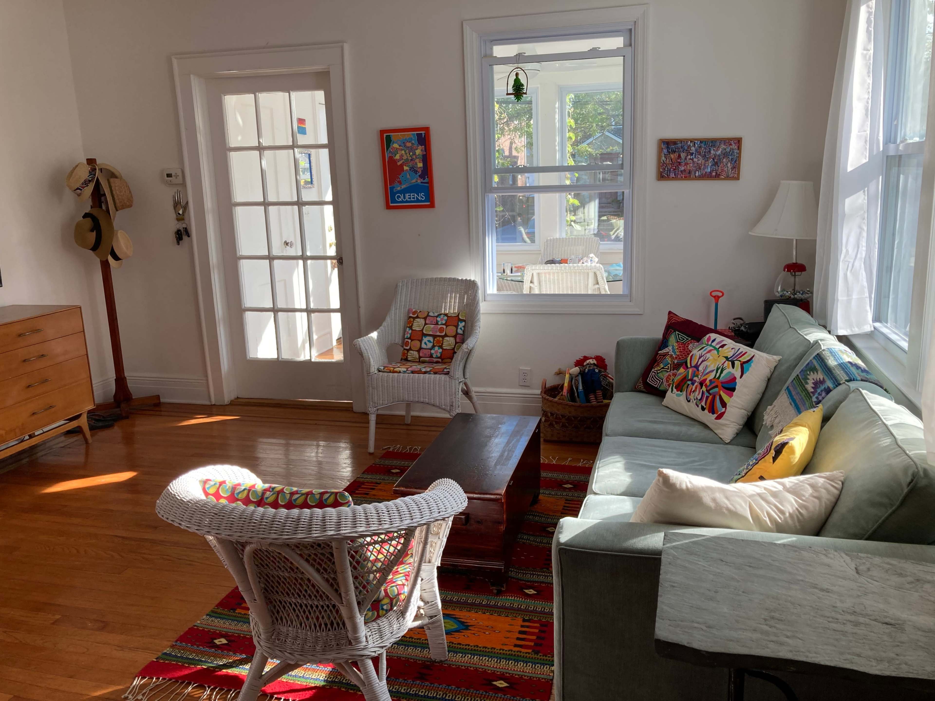 A bright living room featuring a light green sofa, a wooden coffee table, a patterned rug, and a white wicker chair, with sunlight streaming through large windows.