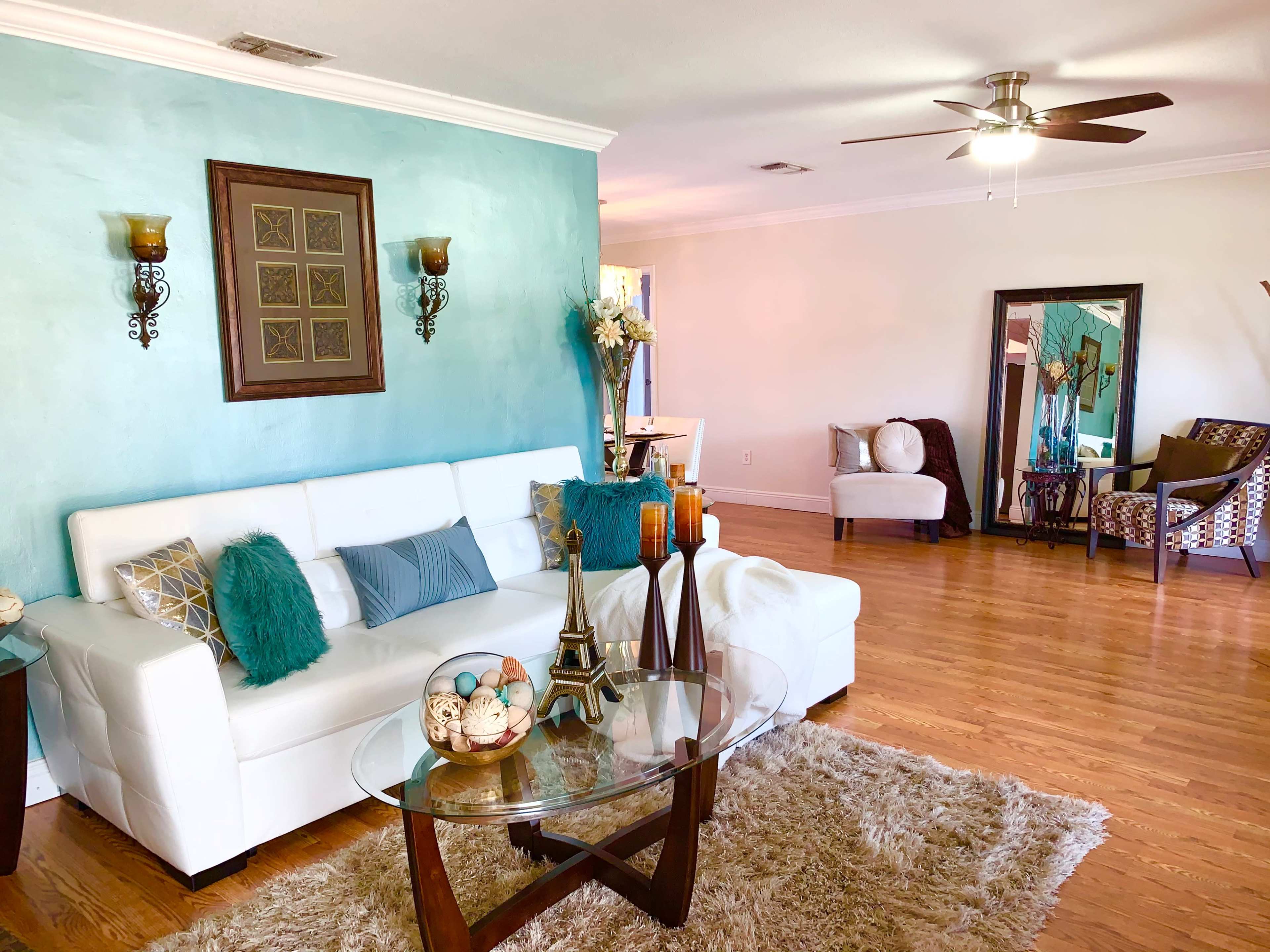 The image shows a modern living room featuring a white sofa, a glass coffee table, and decorative elements, set against a turquoise wall.
