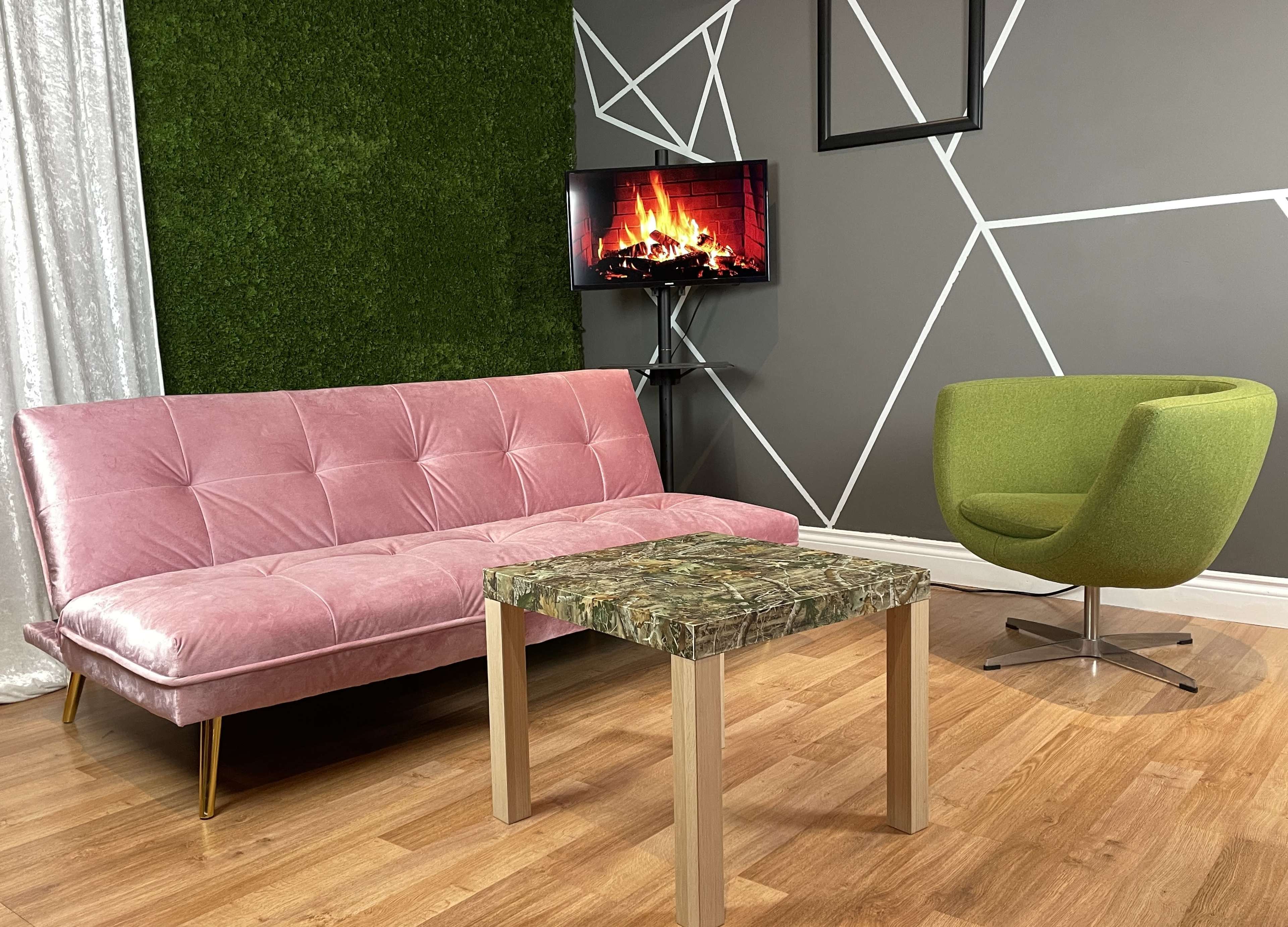 A modern living room setup featuring a pink couch, a green armchair, a marble-patterned table, and a television displaying a fireplace, all against a geometric-patterned wall.