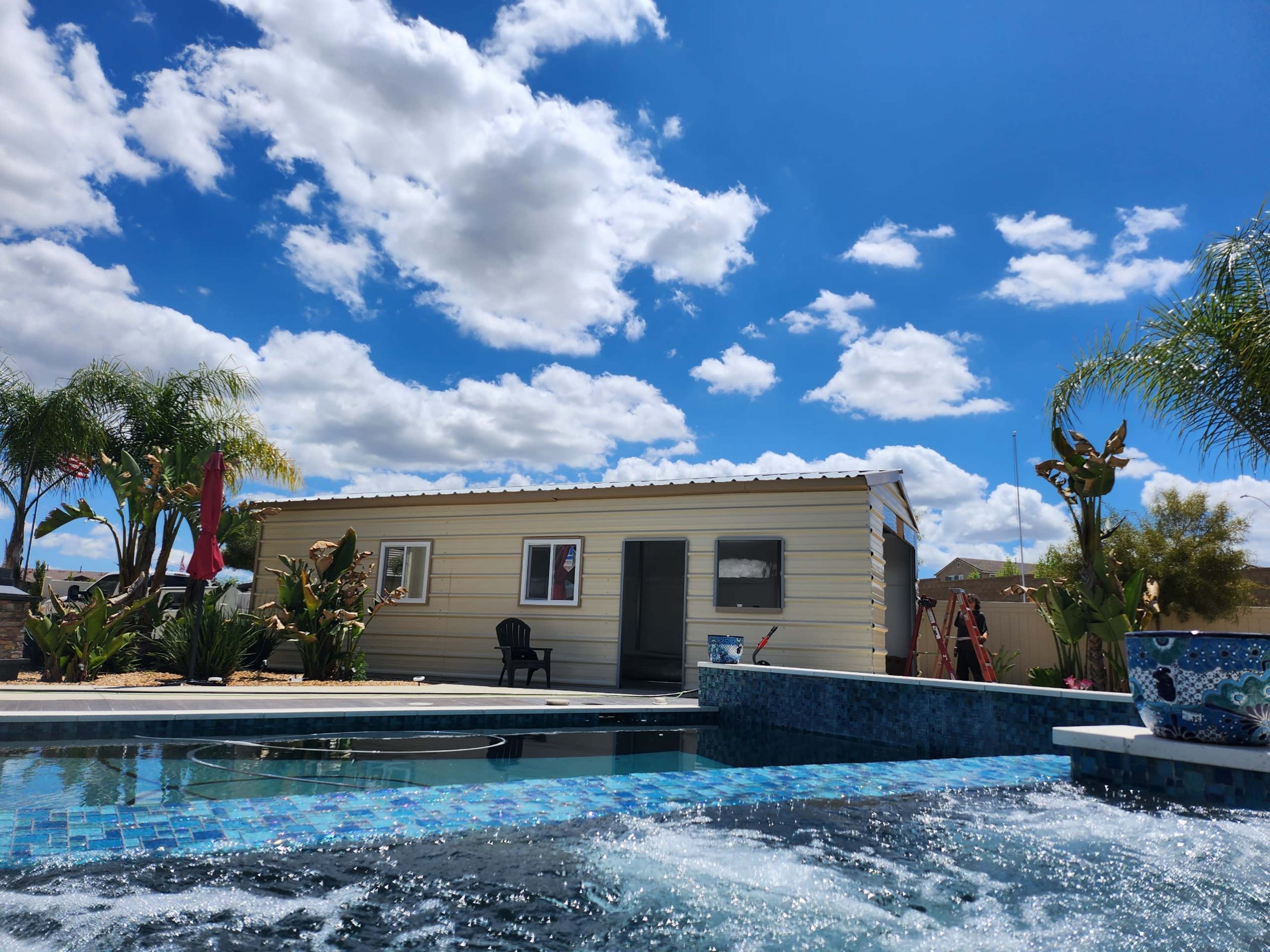 A small metal building is situated beside a pool with blue tiles and palm trees under a partly cloudy sky.