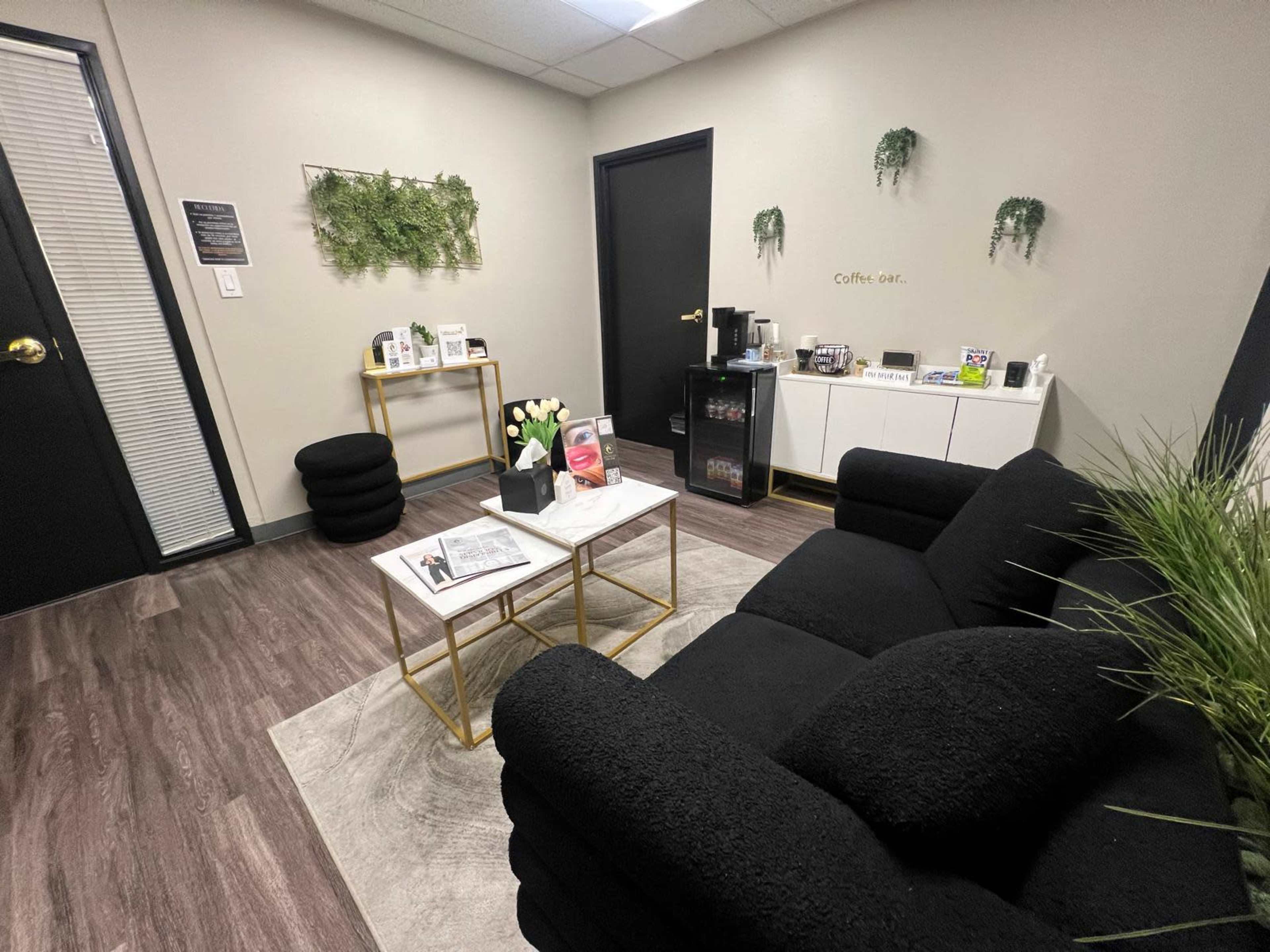 The image shows a modern waiting room with a black sofa, a coffee table, a small table with plants, and a cabinet, all arranged on a wood laminate floor.