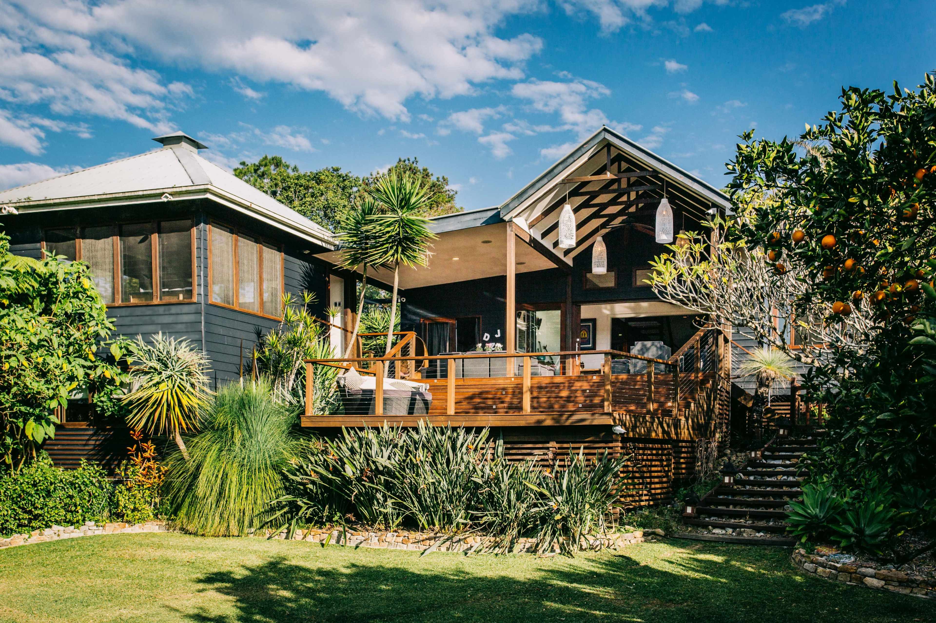 The image shows a two-story wooden house surrounded by lush greenery and a landscaped garden, with a wooden deck and large windows overlooking the yard.