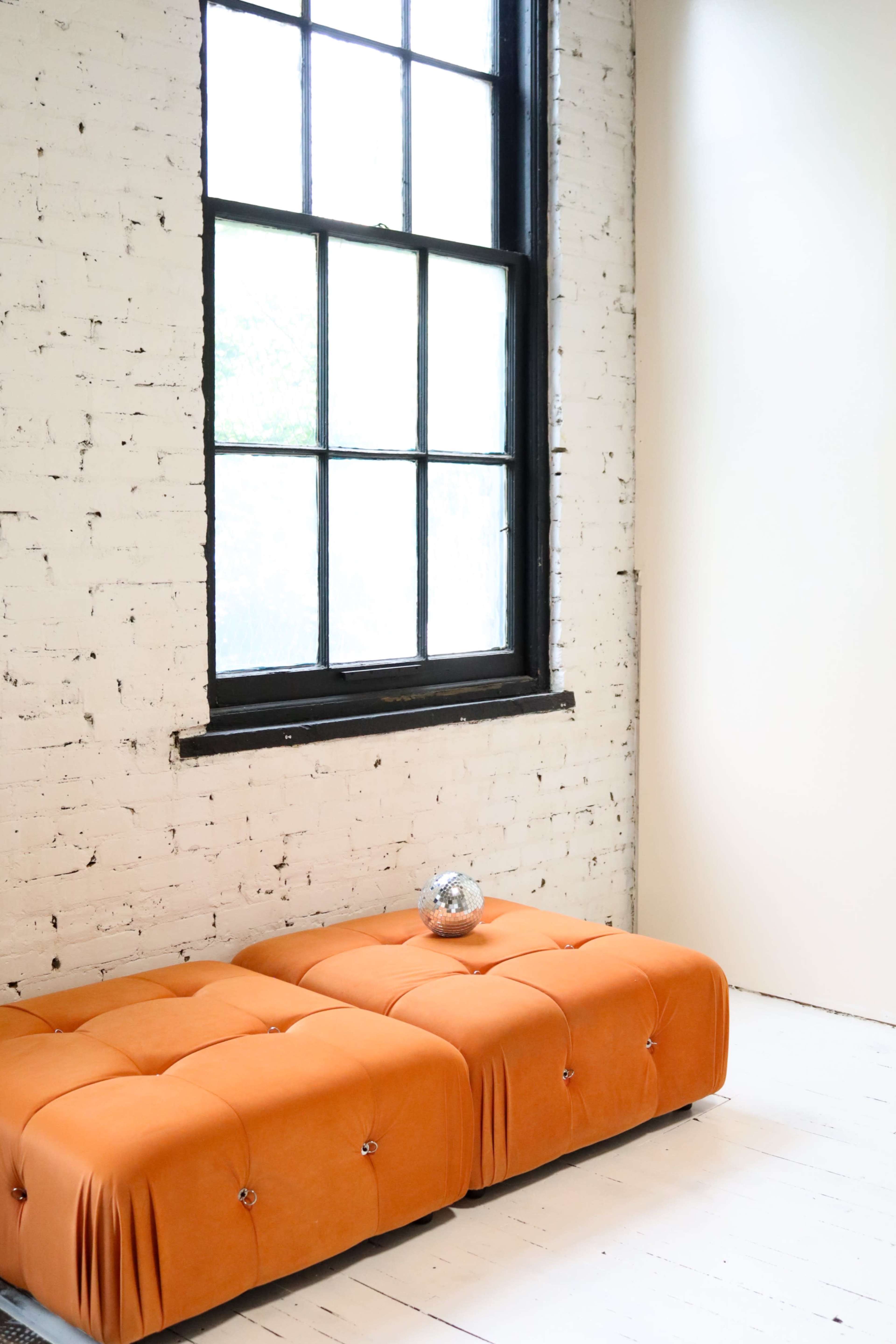 An orange tufted sofa sits against a white brick wall beside a large window with black frames.
