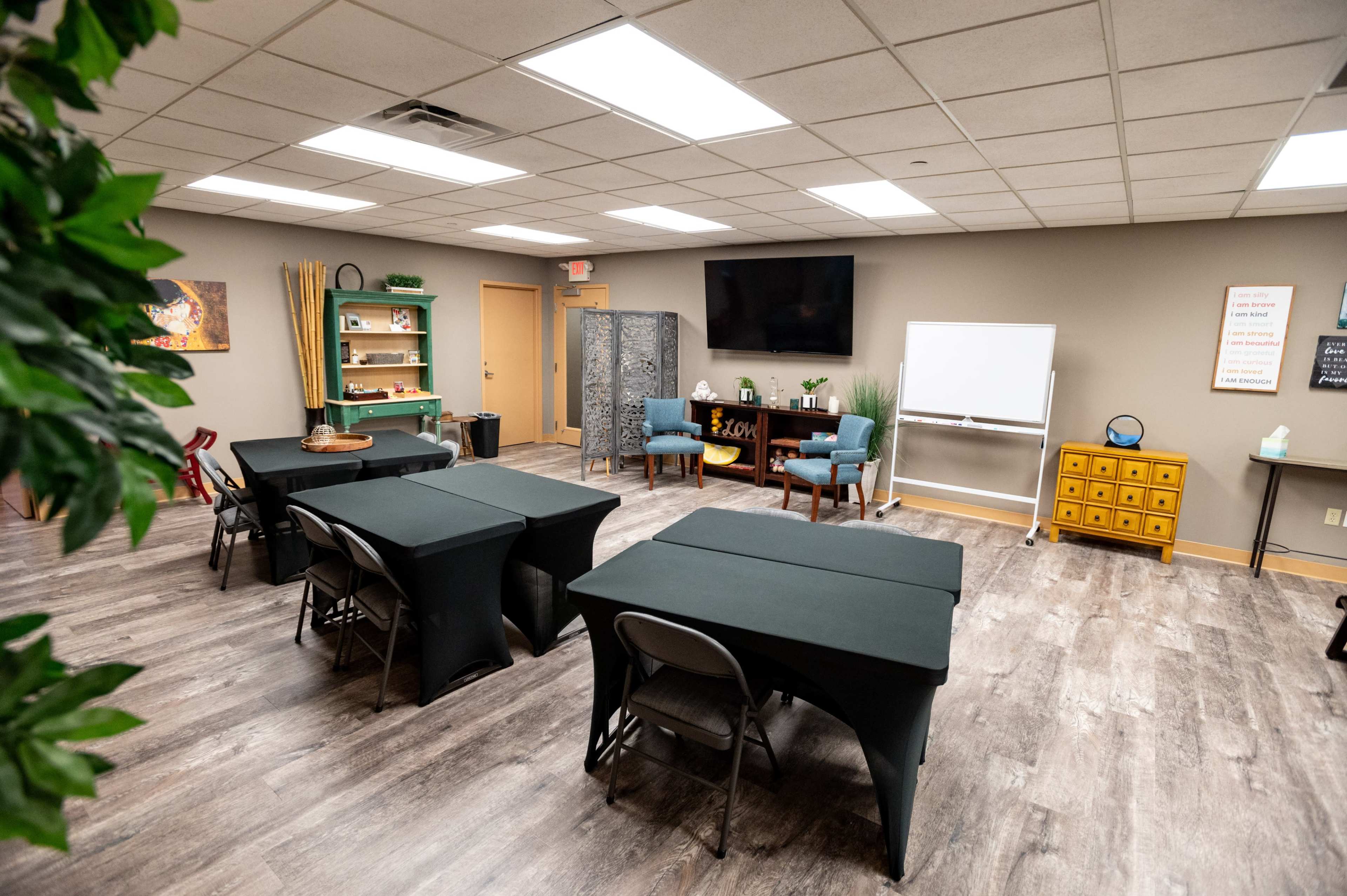 A multipurpose room arranged with black tables, chairs, and a television, featuring a decor of shelving and a whiteboard.