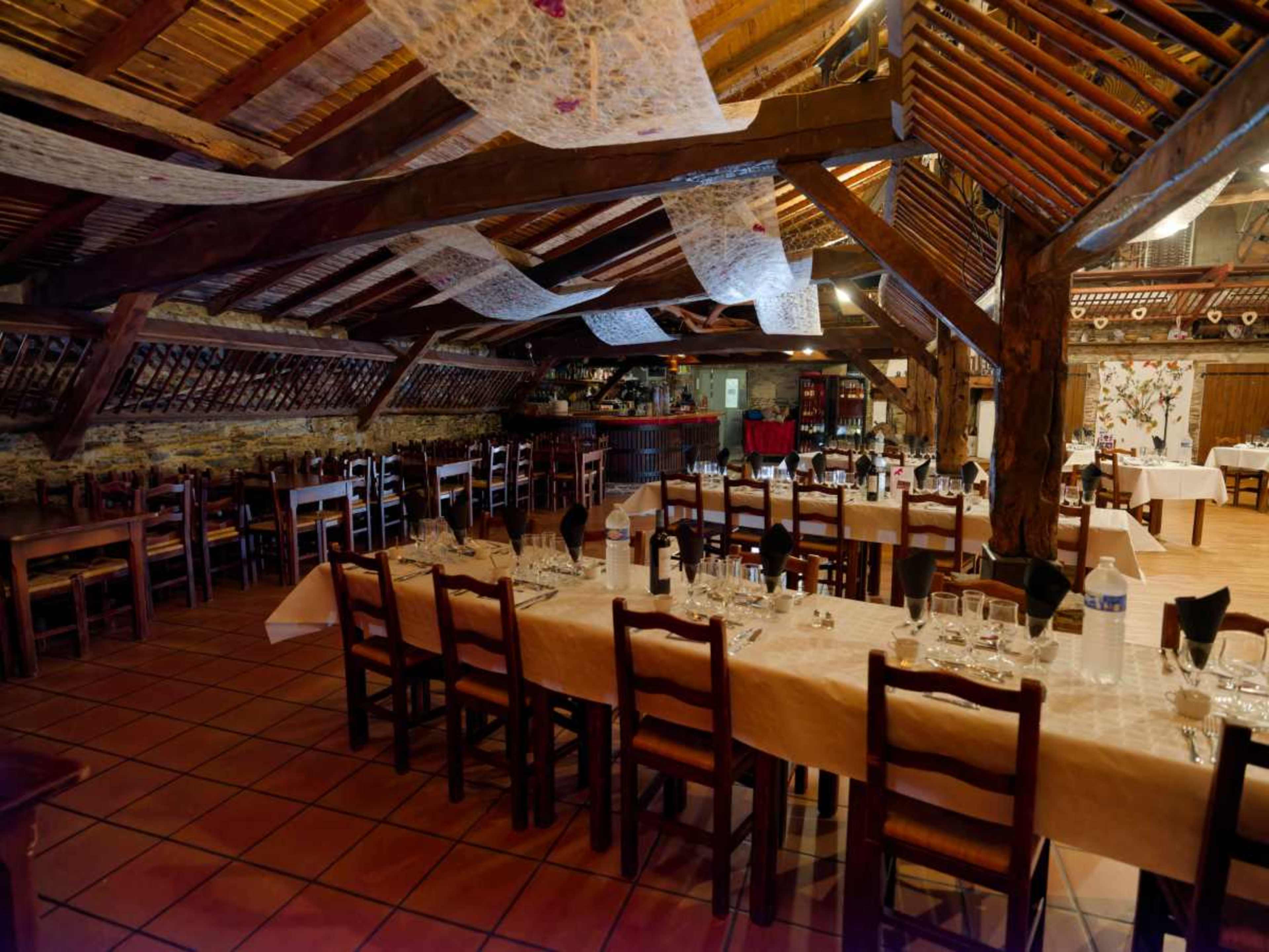 The image shows a spacious restaurant interior with wooden beams, long tables set with white tablecloths, and rustic decor.