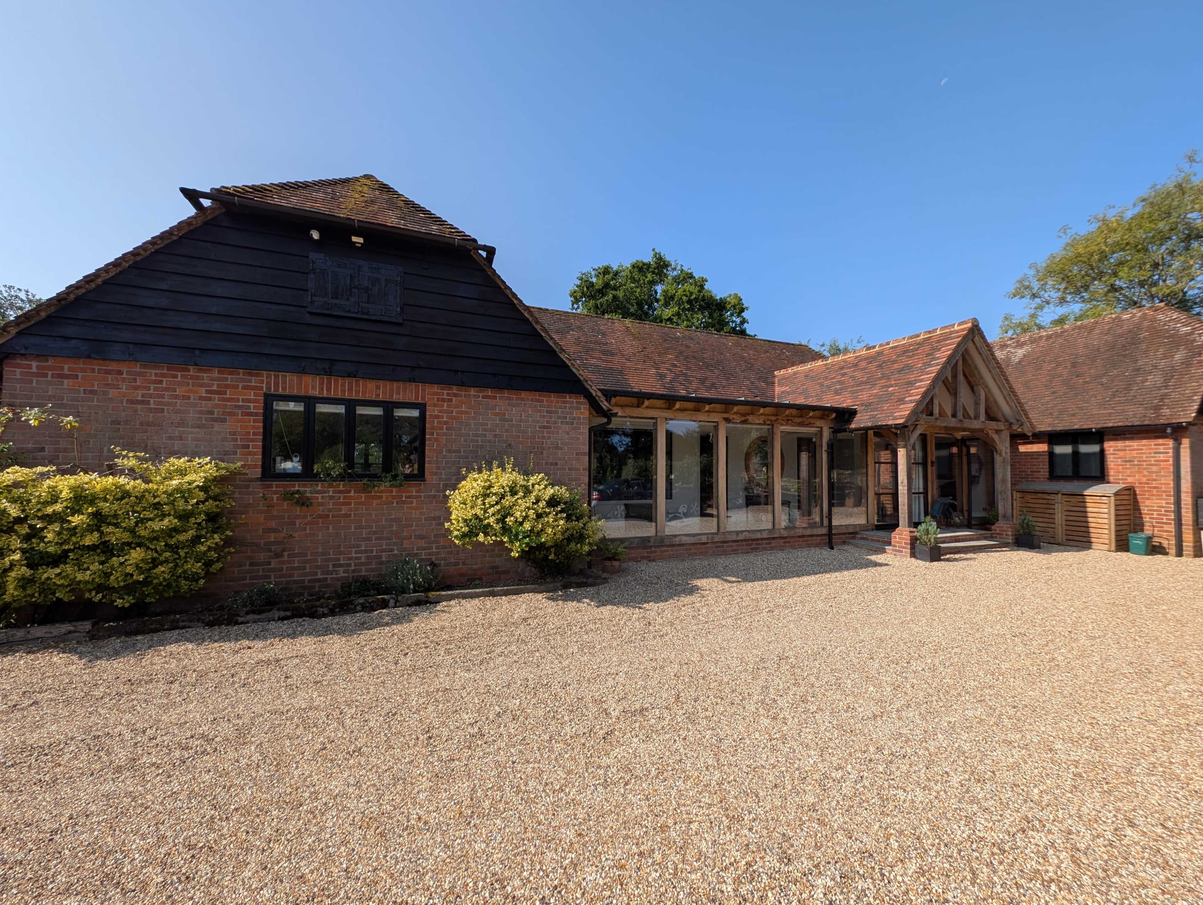 A brick house with a black wooden facade features large glass windows and is surrounded by a gravel driveway and greenery.