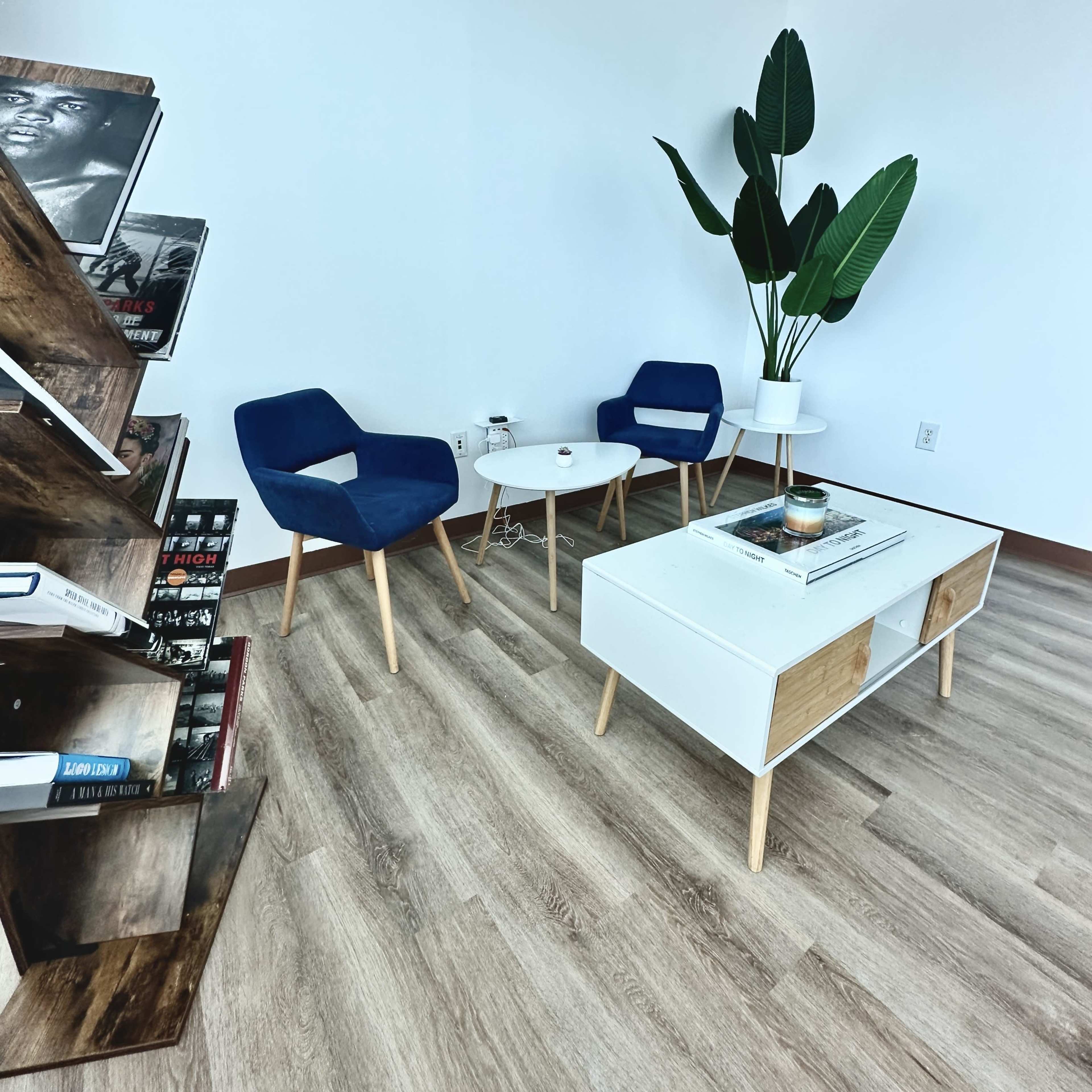 A small reading nook features two blue chairs, a white coffee table, and a tall plant near a bookshelf filled with books.